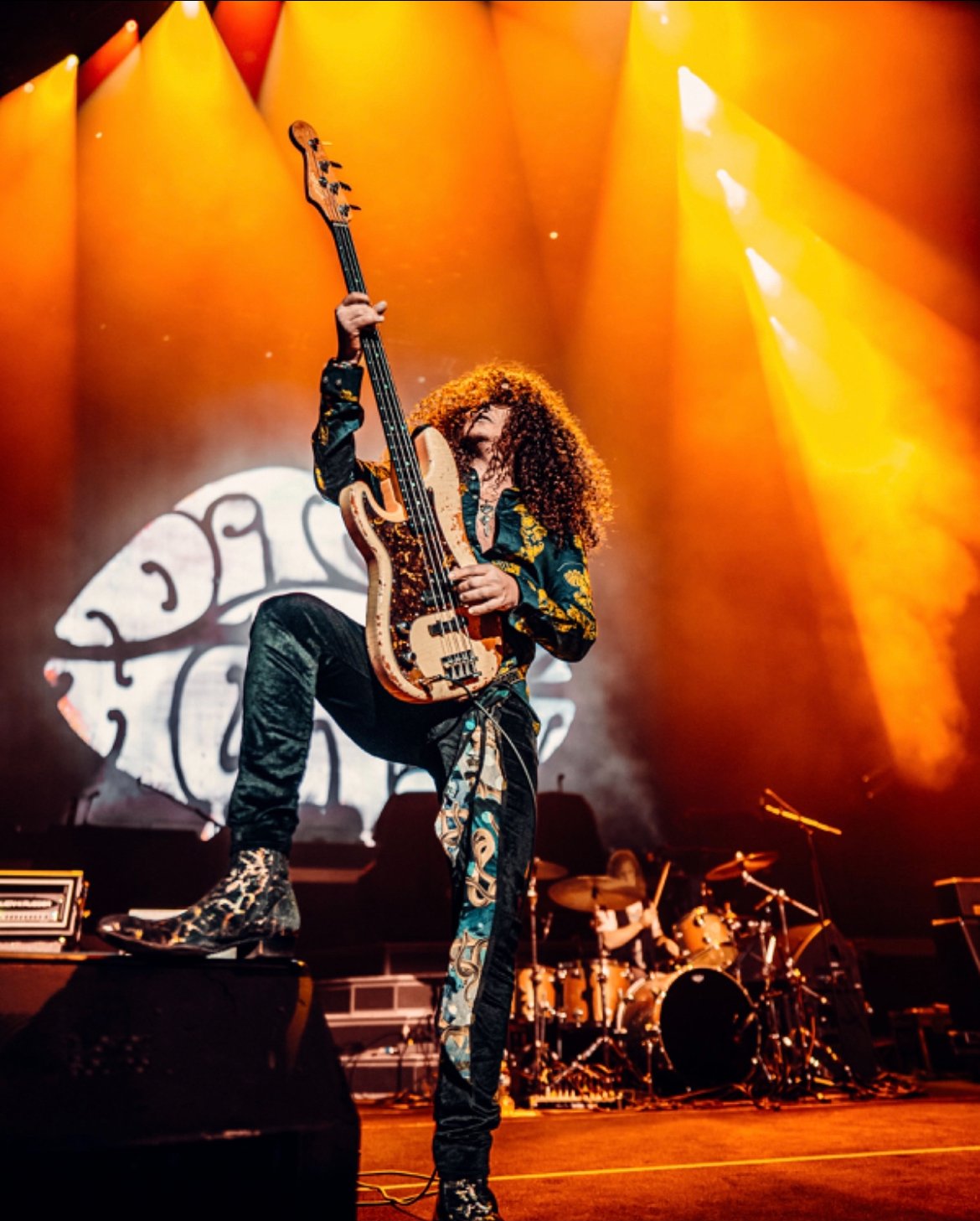 Musician with curly hair playing electric guitar on stage, dramatic lighting, drummer in the background.