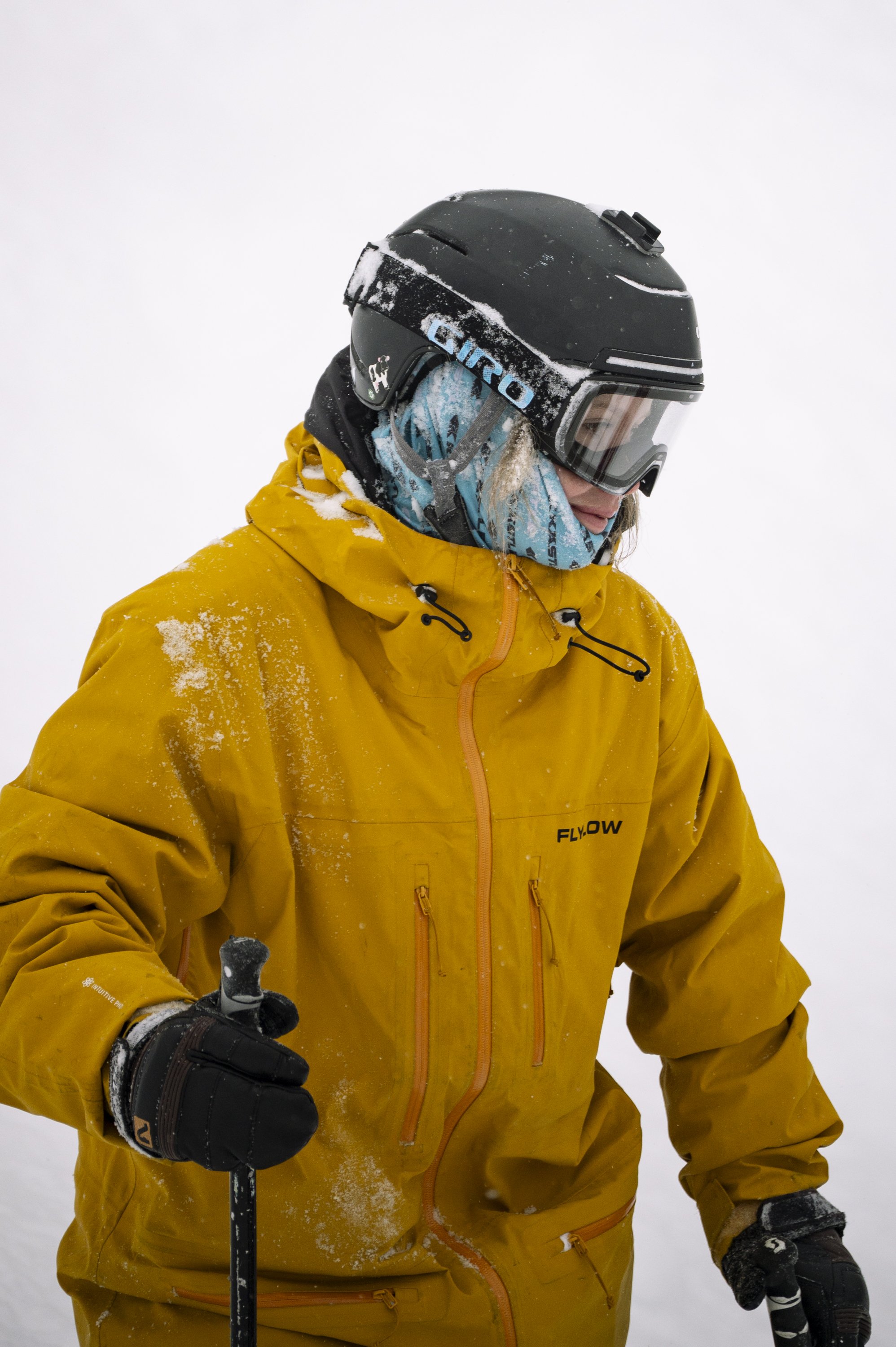 A person dressed in a yellow winter jacket, wearing a black ski helmet with goggles, and holding ski poles, standing in a snowy environment.