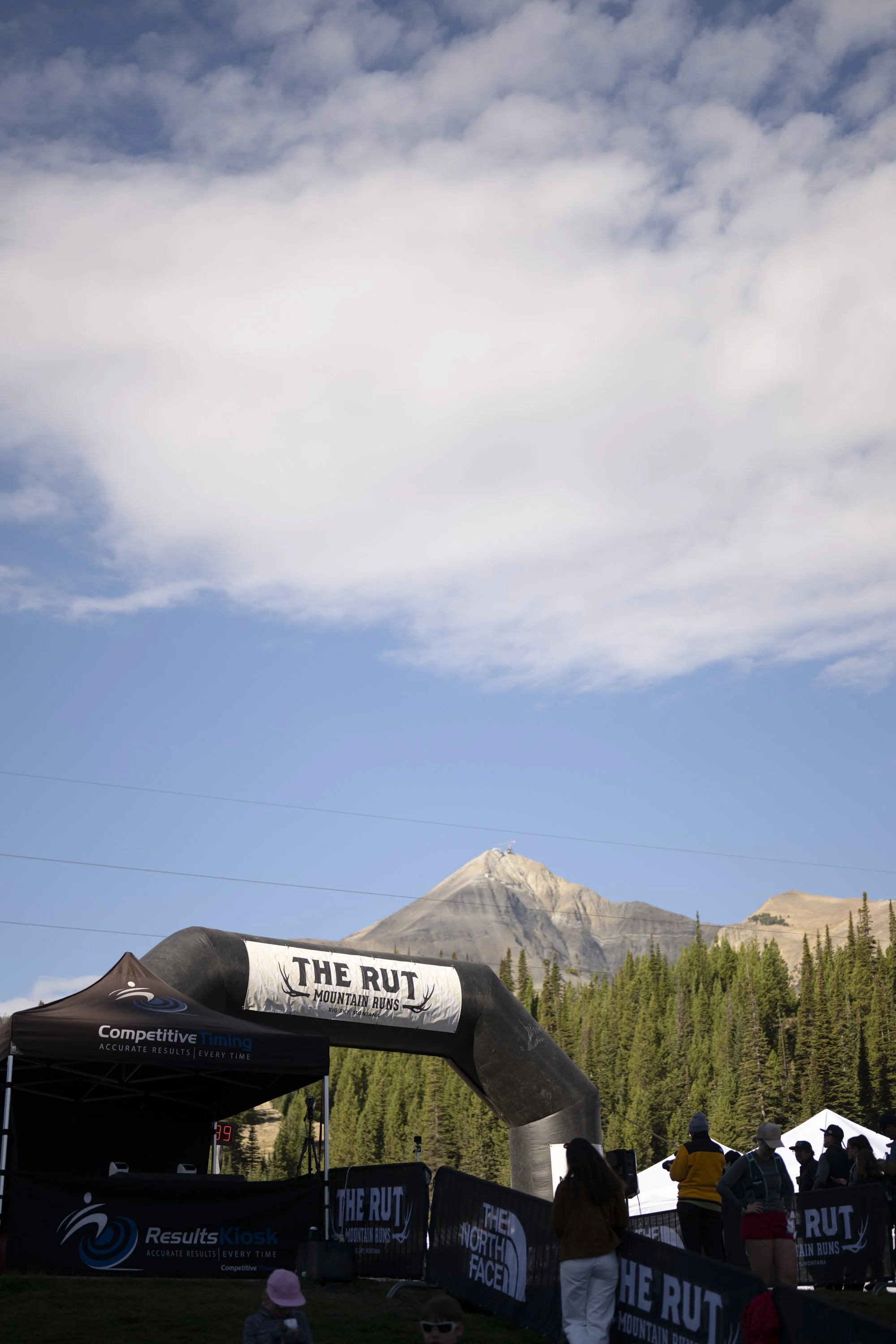 People gathered at a mountain race finish line, with a large inflated arch labeled 'The RUT Mountain Runs' and a mountain in the background under partly cloudy blue sky.