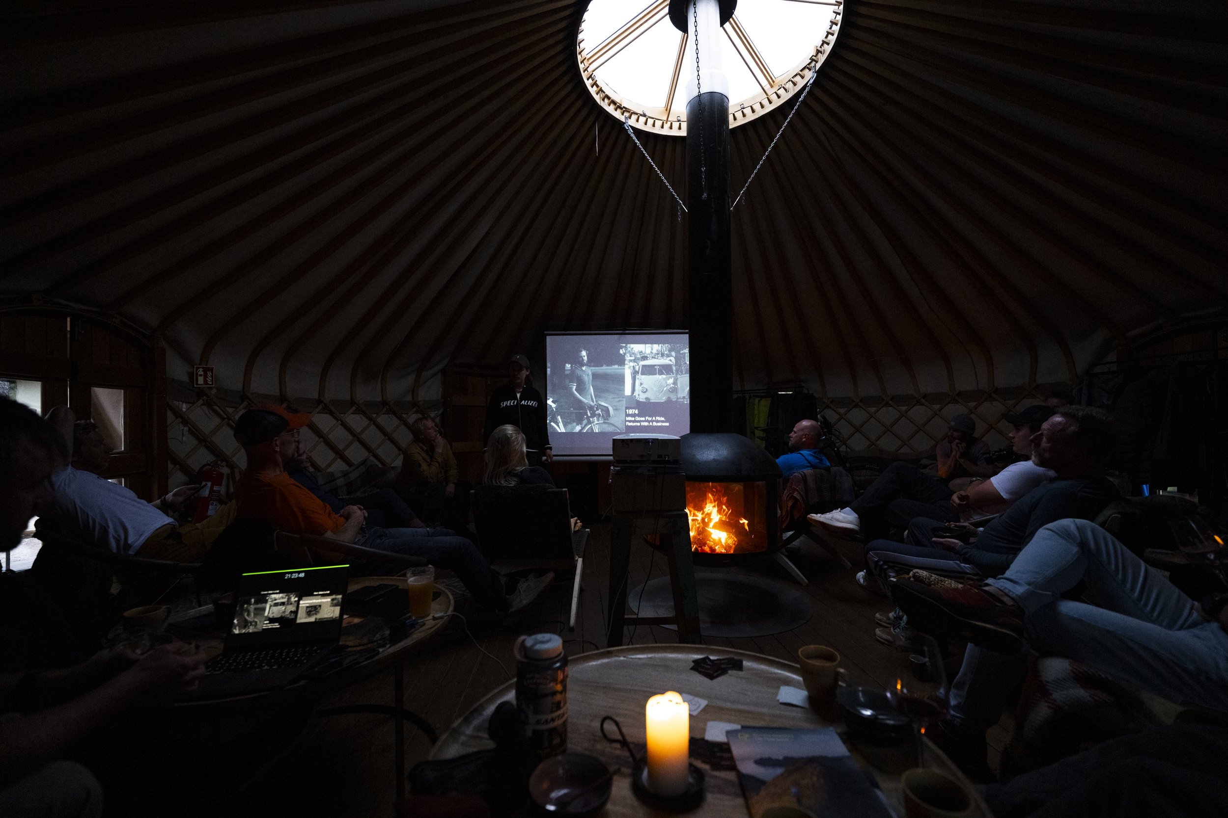 A group of people seated inside a dimly lit, wooden, yurt-shaped structure, watching a presentation with a screen showing black and white photos. There is a chimney with a fire burning, and the ceiling has a large circular opening letting in natural 