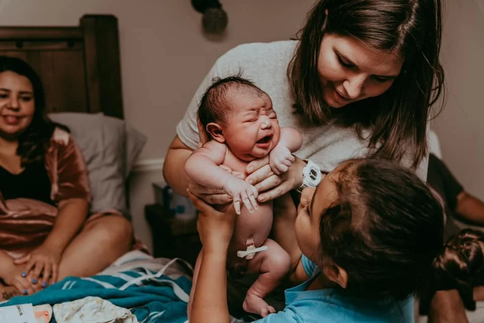 Midwife Amy holding a crying newborn as an older sibling reaches up to greet the baby