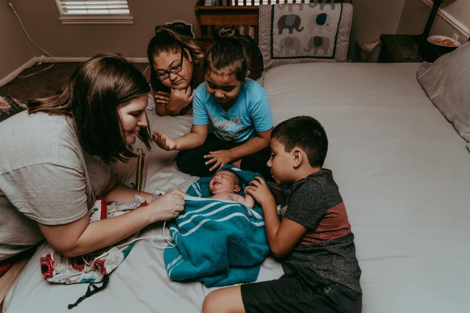 Family gathered on the bed watching the midwife perform a newborn assessment after a home birth