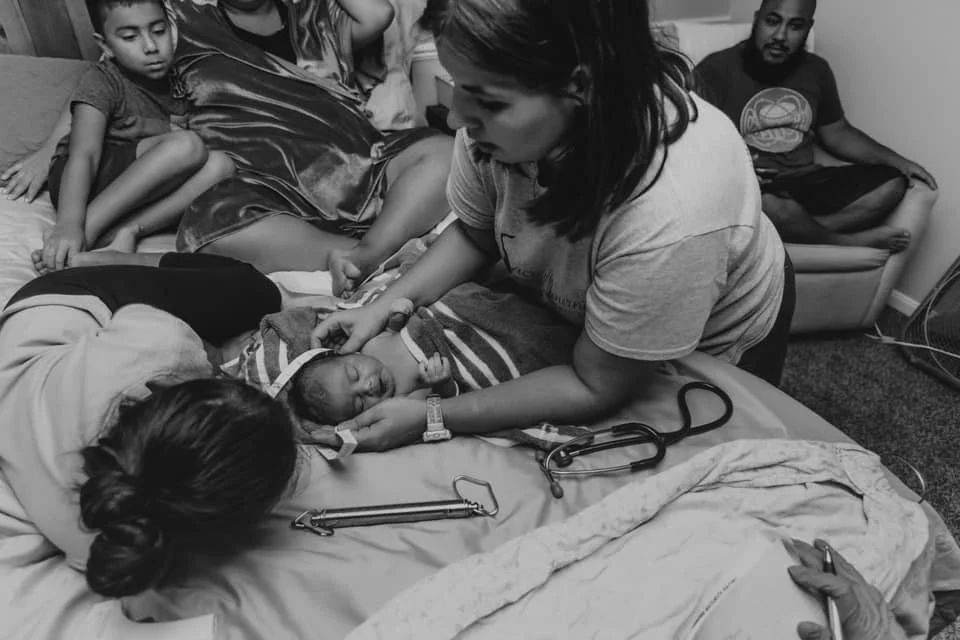 Midwife examining and weighing a newborn on the bed while the family looks on after a home birth