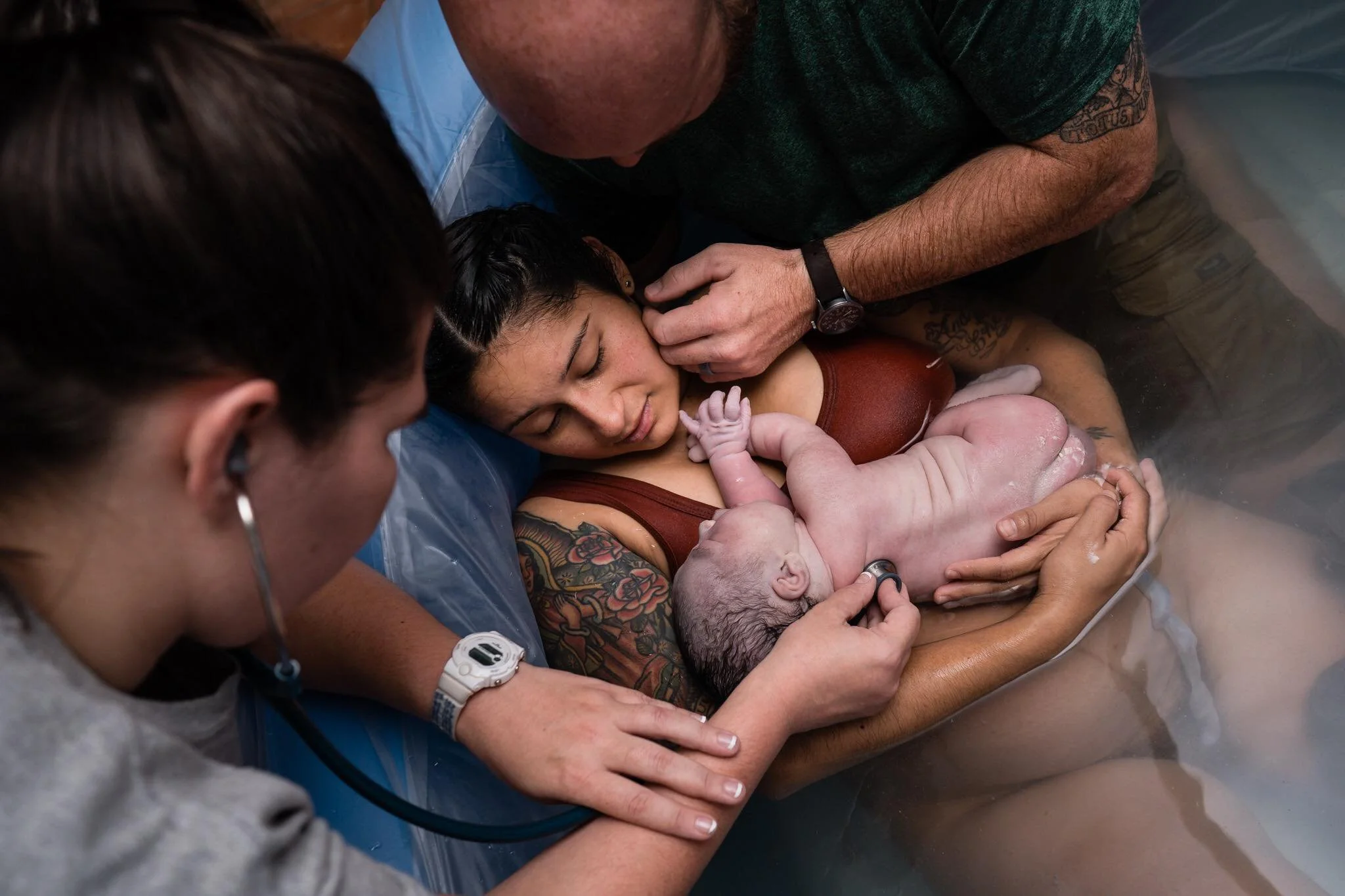 Midwife listening to a newborn's heartbeat with a stethoscope as the mother holds her baby in the birth pool, dad close beside them
