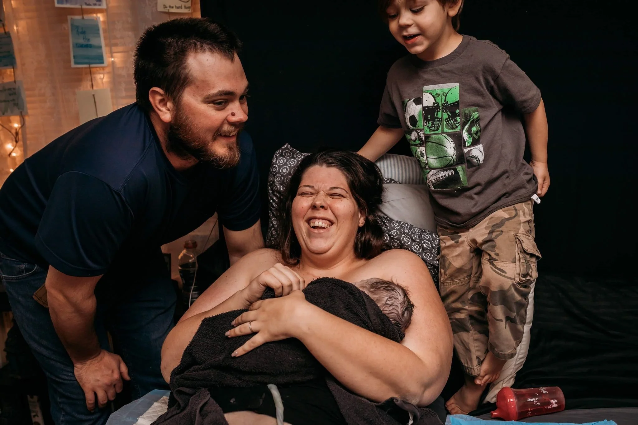 Mother laughing with her husband and son while holding her newborn wrapped in a towel just after a home birth