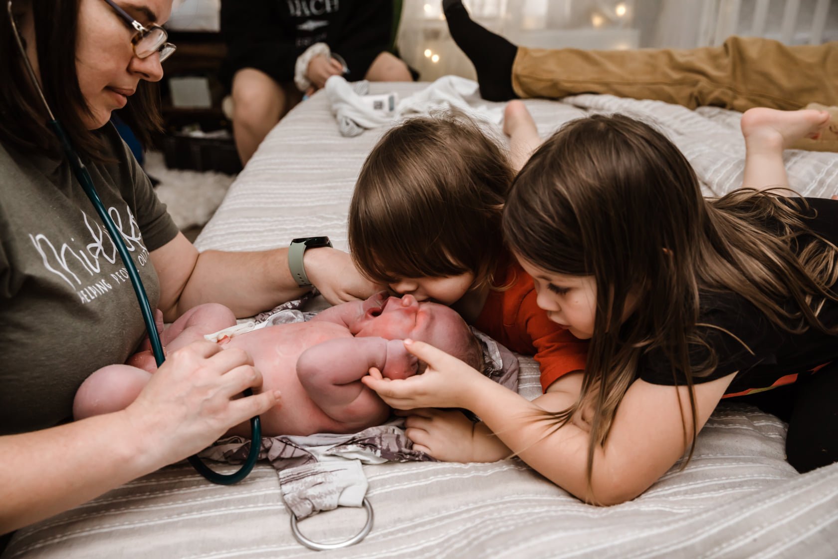 Siblings kissing their newborn baby brother during the midwife's newborn exam at home