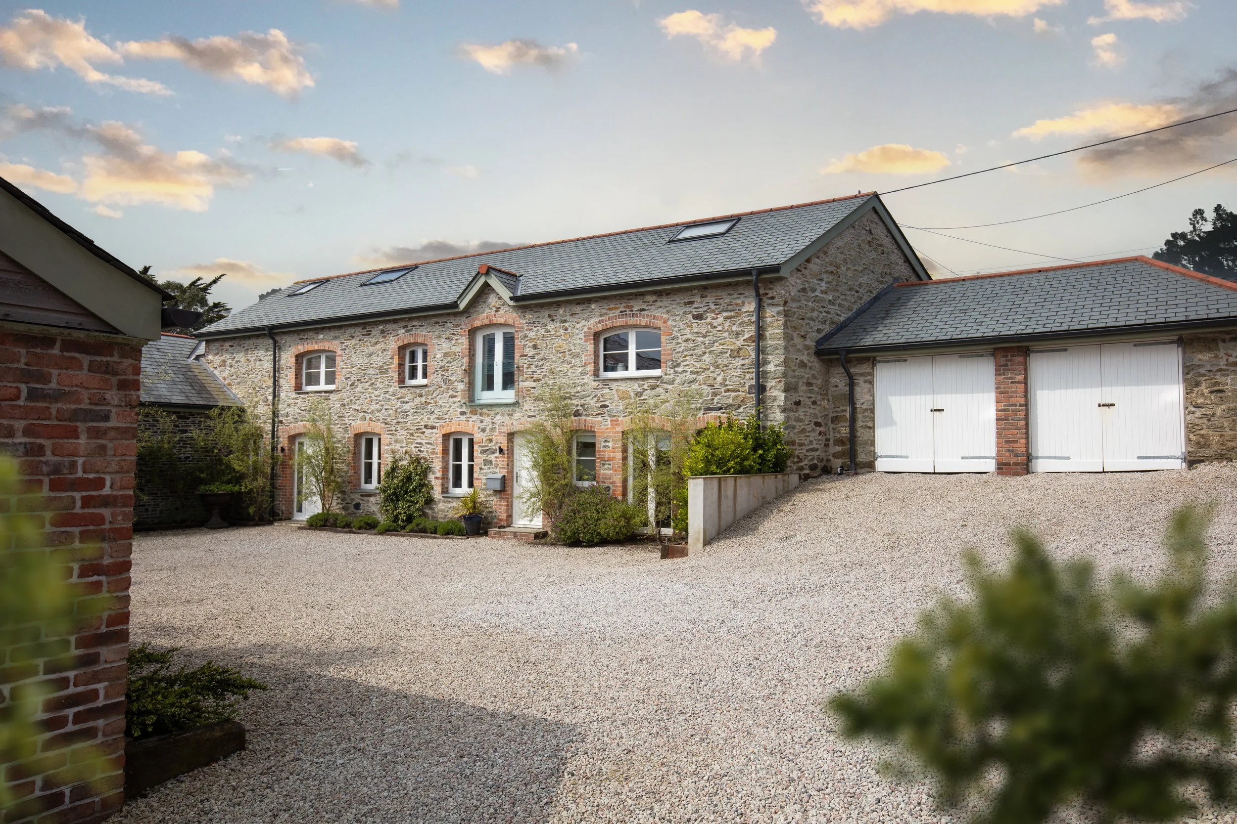 A stone house with a gray roof and multiple windows, surrounded by a gravel driveway and some greenery, under a partly cloudy sky.