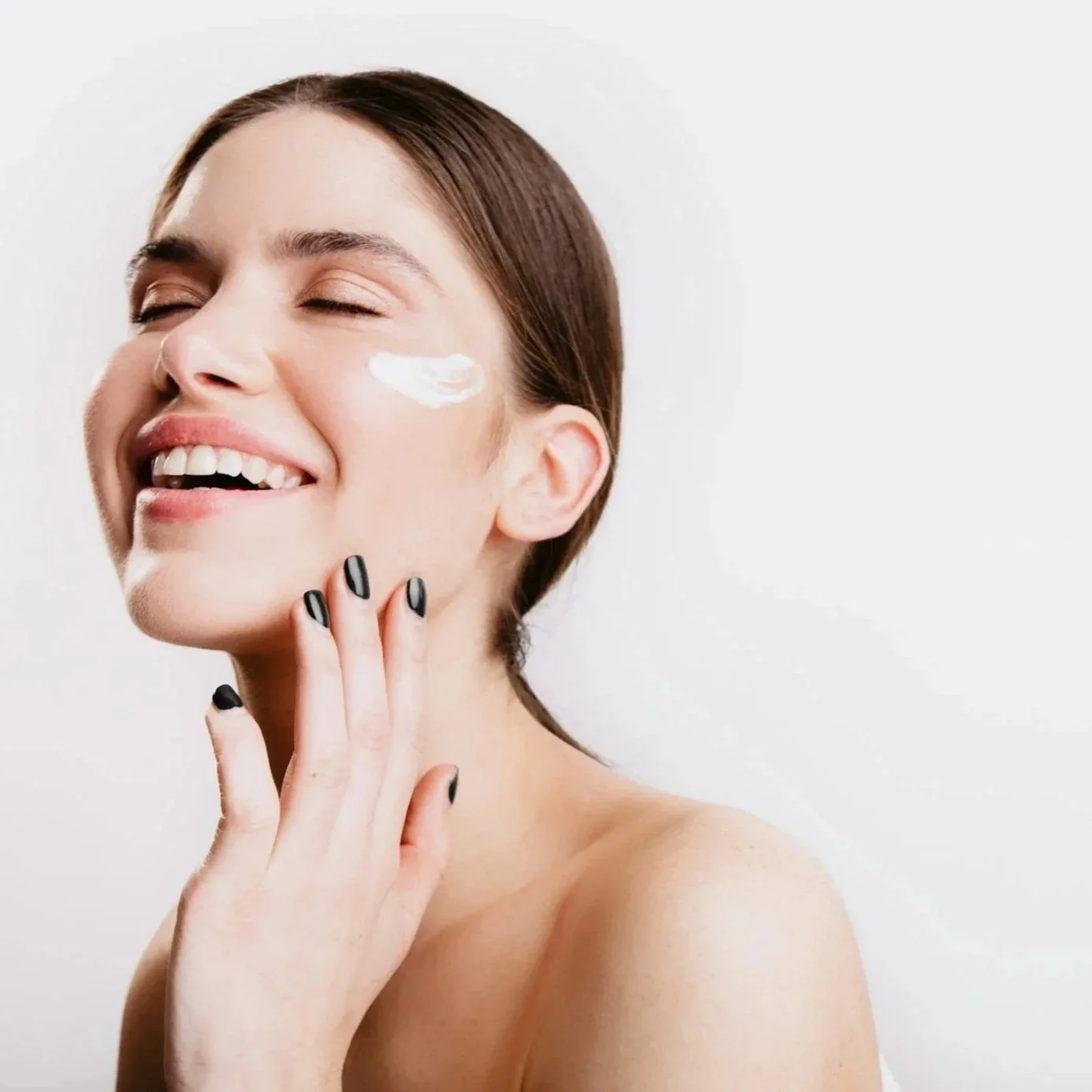 A woman with brown hair applying moisturizer to her face, smiling, with black nail polish on her fingers, against a white background.