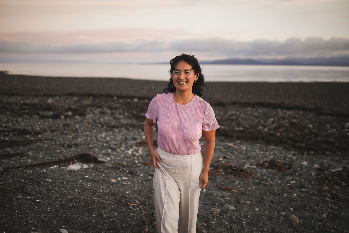 A woman smiling and posing on a rocky beach at sunset, wearing a pink shirt and light-colored pants.