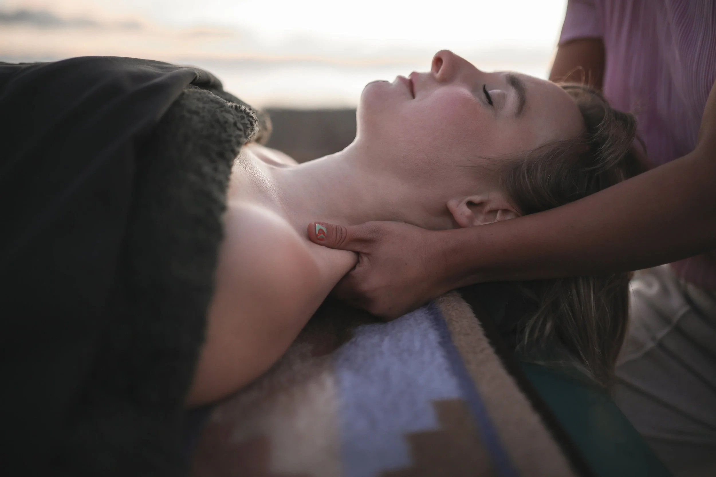 A young woman lying down with her eyes closed while a person gently holds her neck and shoulder. The woman has long light brown hair, fair skin, and is wearing a purple shirt. The background is blurred, indicating an outdoor setting with a sunset sky.