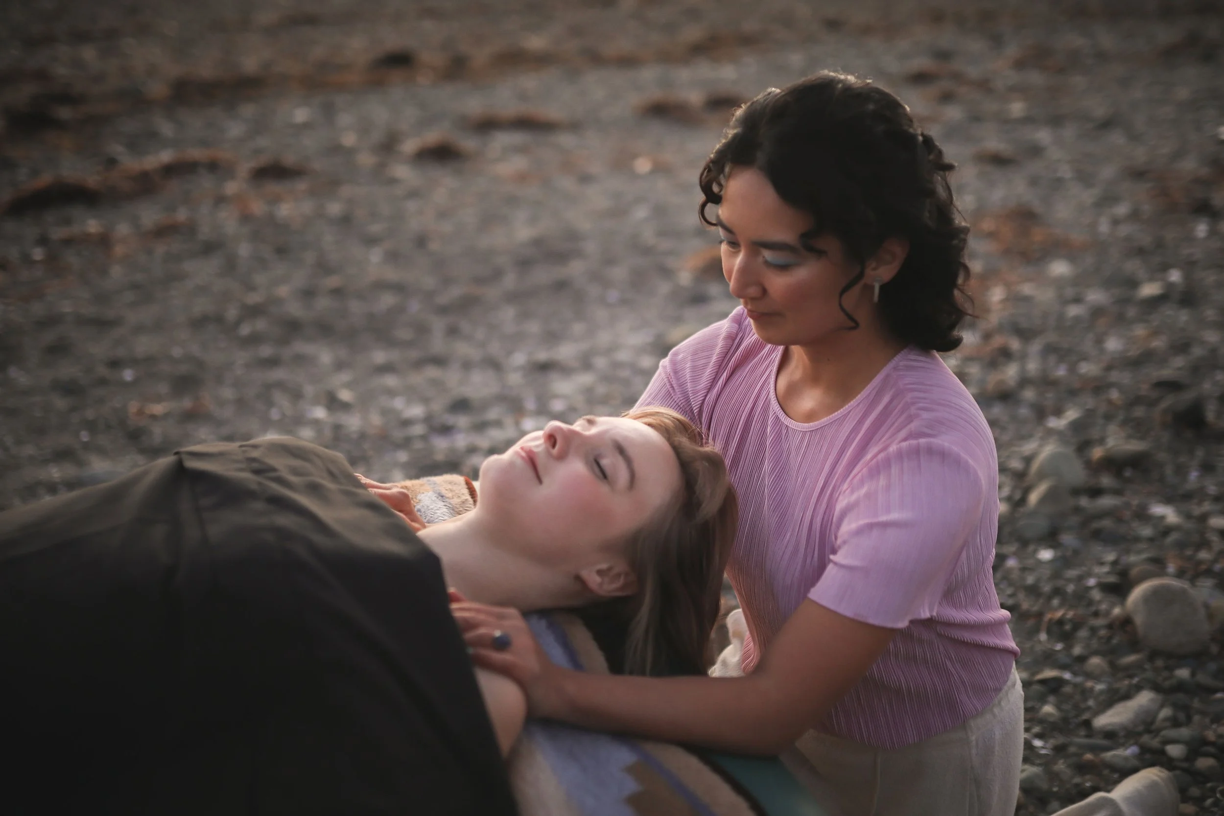 A woman wearing a pink shirt comforts a person lying on rocky ground, holding their head gently, at sunset or dusk.