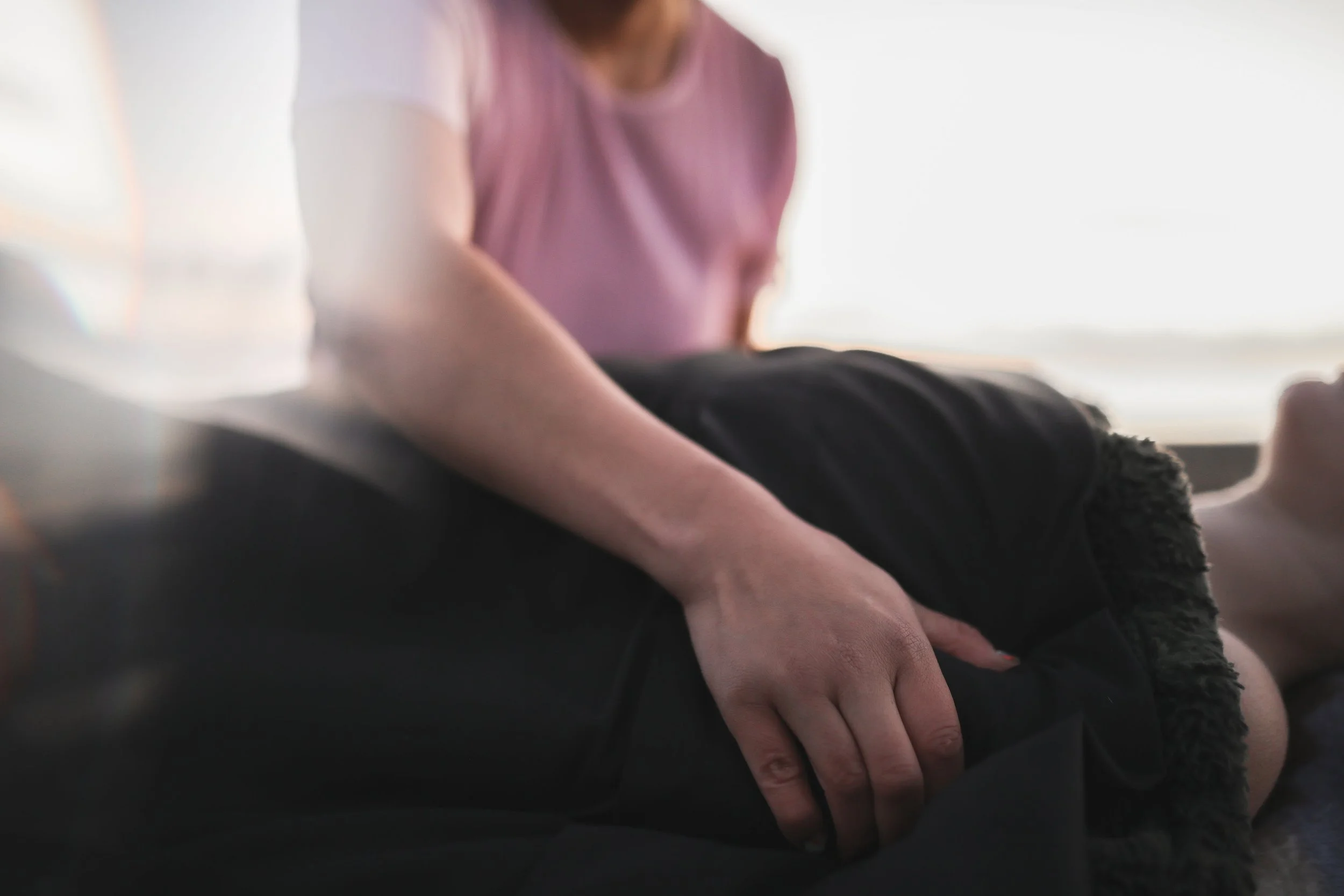 A person with light skin wearing a pink top and dark pants sitting on a dark rug, with one arm resting on their knee, in a bright room with soft lighting.