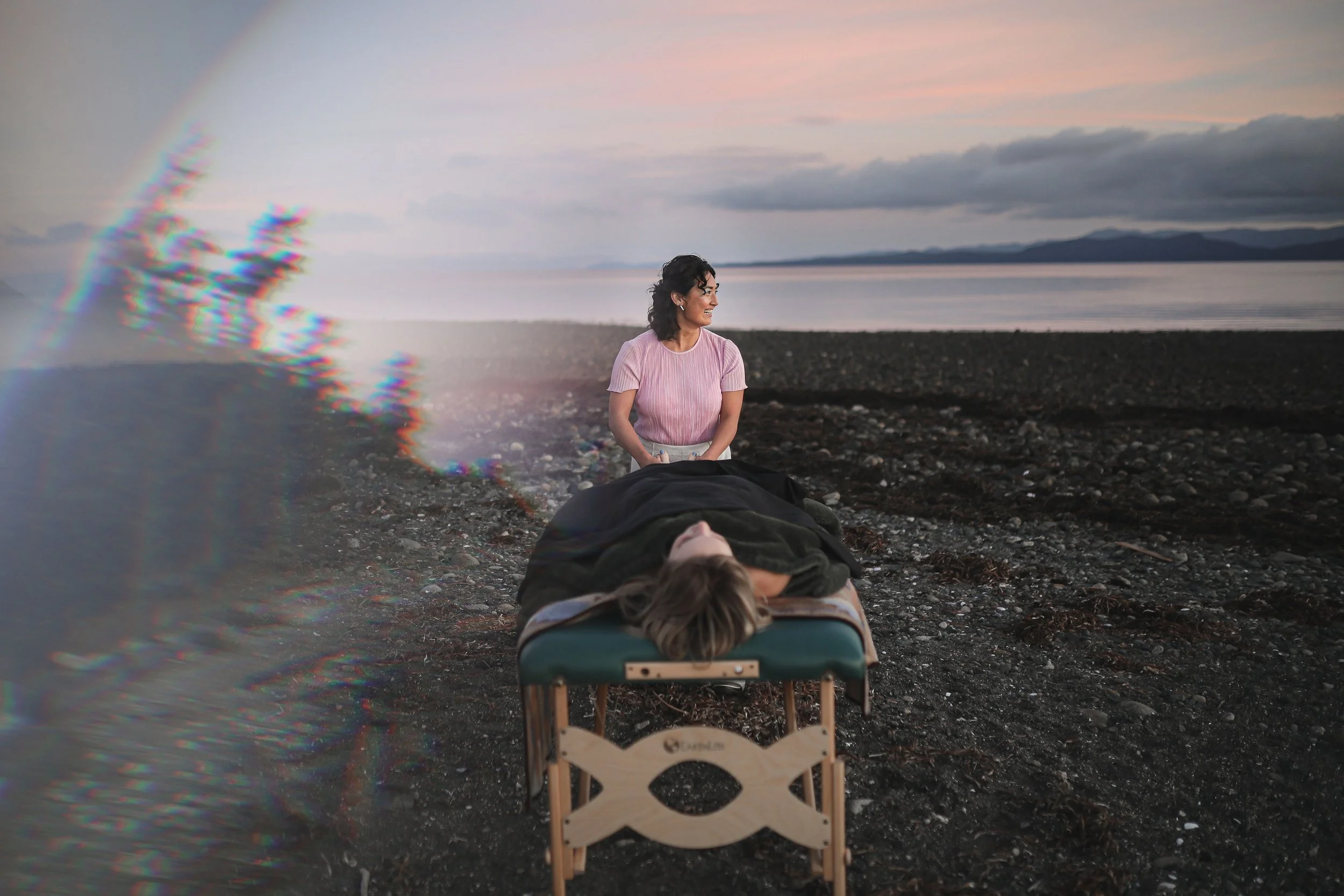 A woman sitting on a rocky beach at sunset, smiling and looking to the side. Two people are lying face down on a small wooden massage table in front of her. The scene includes a rainbow on the left side and distant mountains across a body of water under a partly cloudy sky.