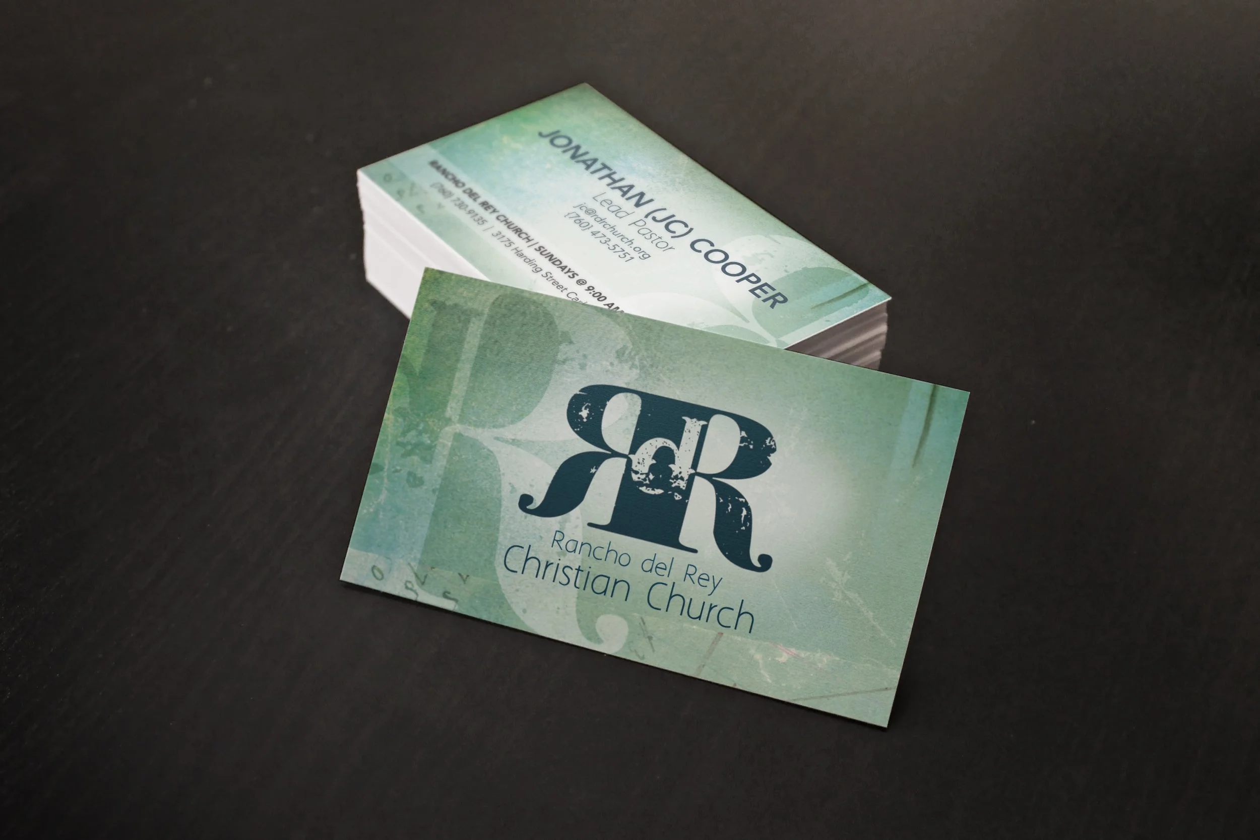 Business card design for church, Rancho del Rey Christian Church, featuring a green watercolor background, a large stylized 'R' with a decorative design, and contact information on one card, with the church's name and logo on the other card.