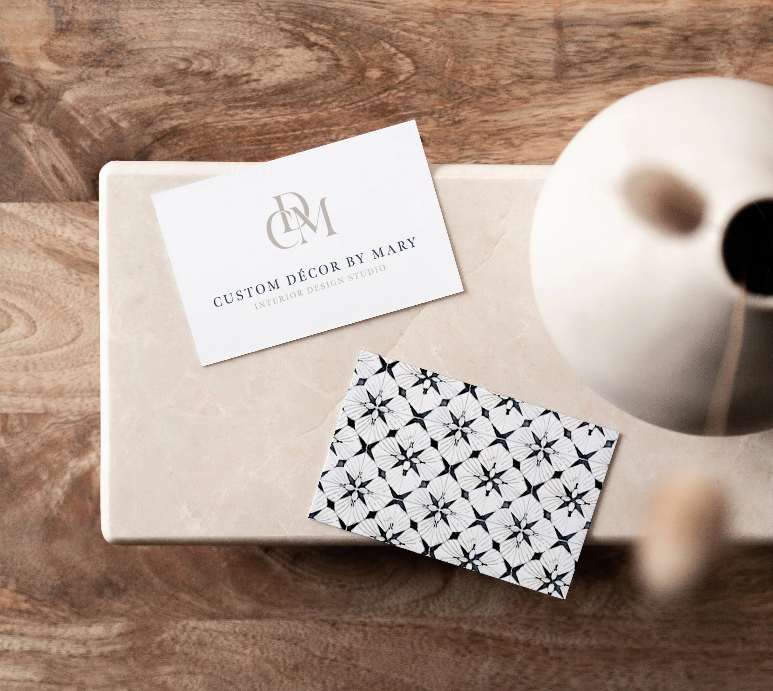 Business card design for interior designer, San Diego CA, Custom Décor by Mary, interior design studio, on a beige marble surface, next to a black and white patterned card, and a white vase with black accents.