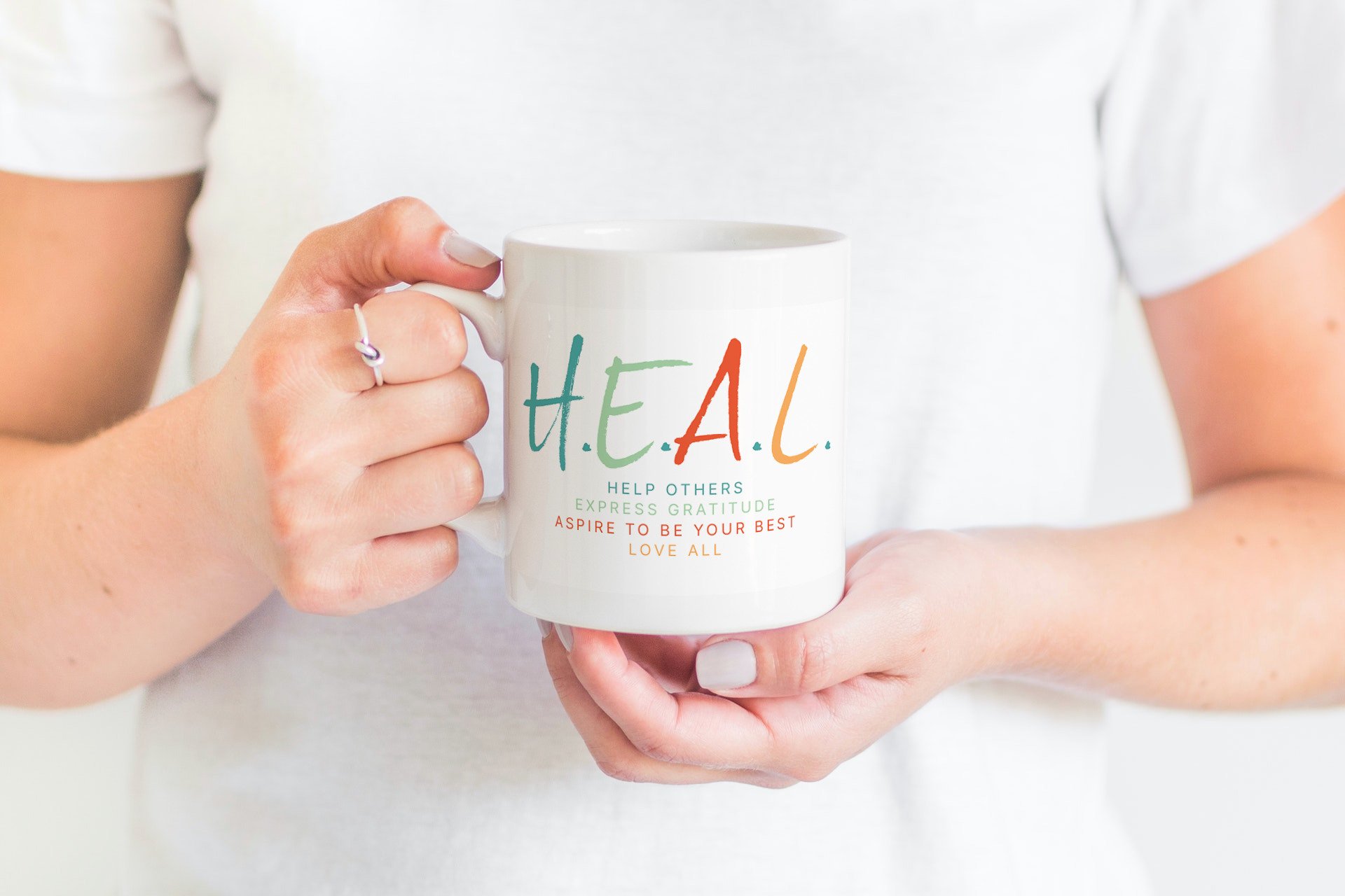 Person holding a white mug with mantra design for shop and merch items, the word 'H.E.A.L.' and motivational phrases about gratitude and love.