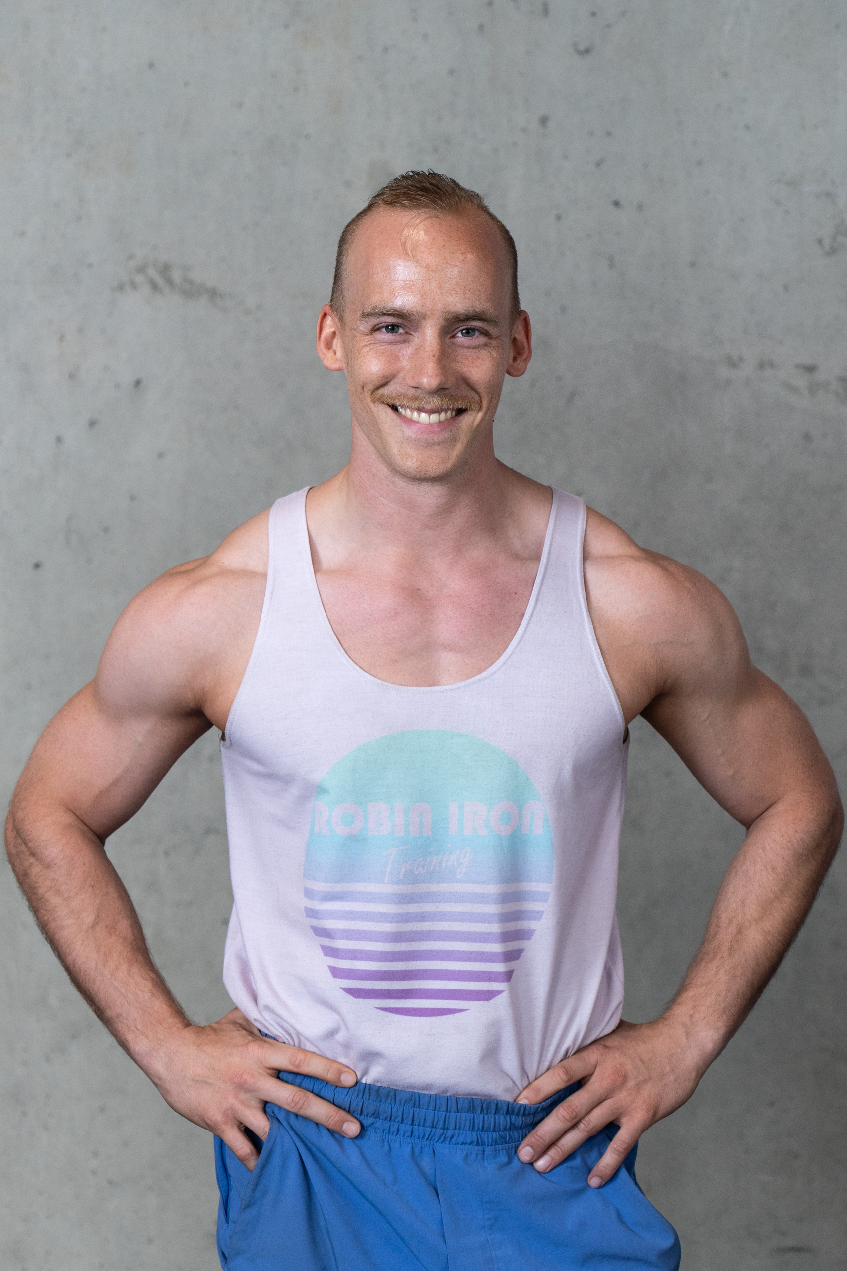A muscular man with short hair and a mustache smiling, wearing a white tank top and blue shorts, standing with hands on hips in front of a gray concrete wall.