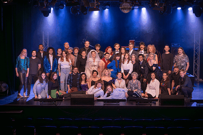 Group of musical theatre performers on stage with colourful lighting, some dressed in formal attire, posing for a photo during or after a musical theatre performance or event.