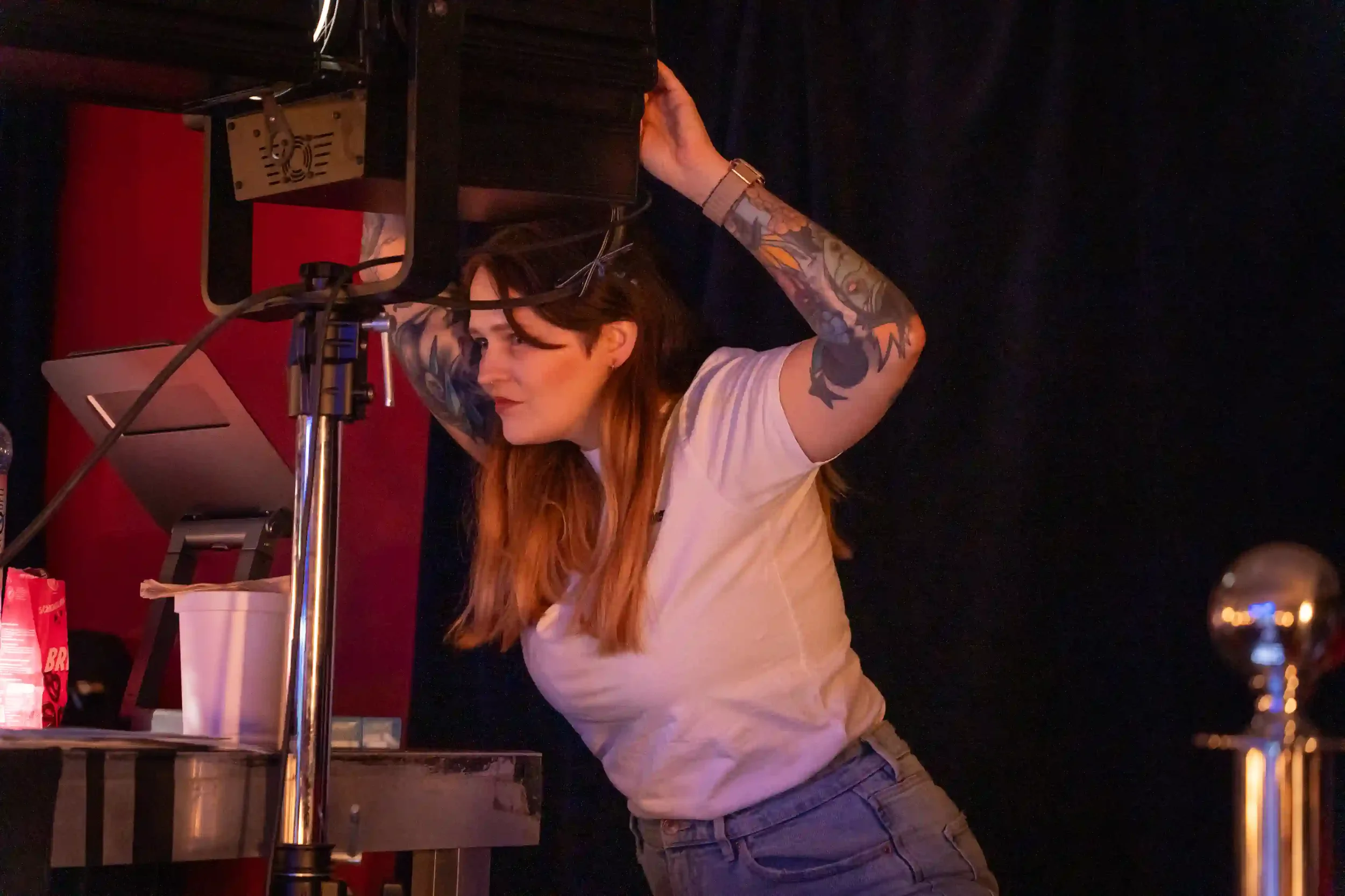 A woman with long hair and tattoos is adjusting a spotlight in a dark theatre with a red wall and black curtains.