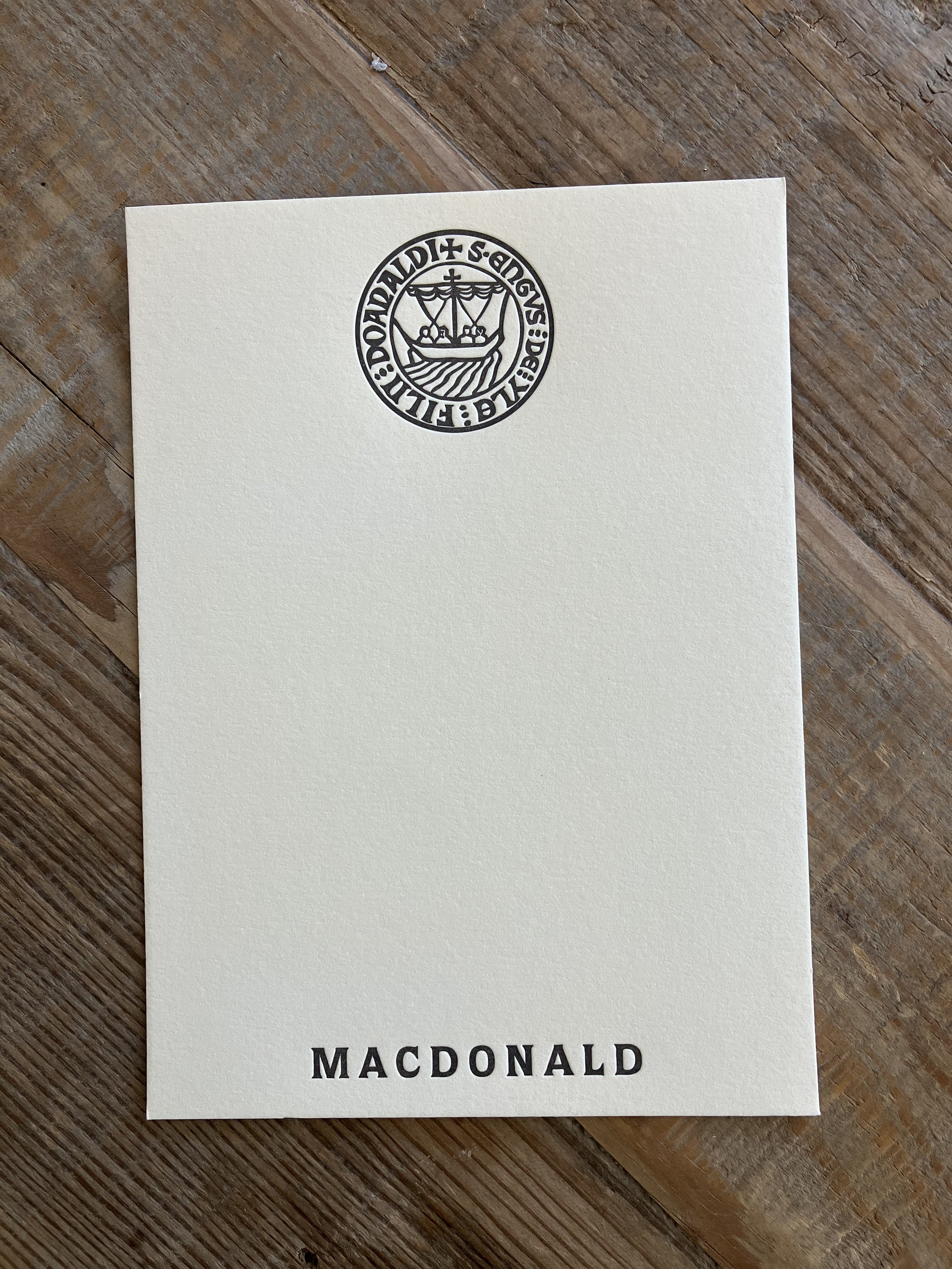 A piece of cream-colored stationery with "MACDONALD" printed at the bottom and a circular emblem featuring a heraldic ship and the words "S: DONALD + FILII A. MACOE" at the top, placed on a wooden surface.
