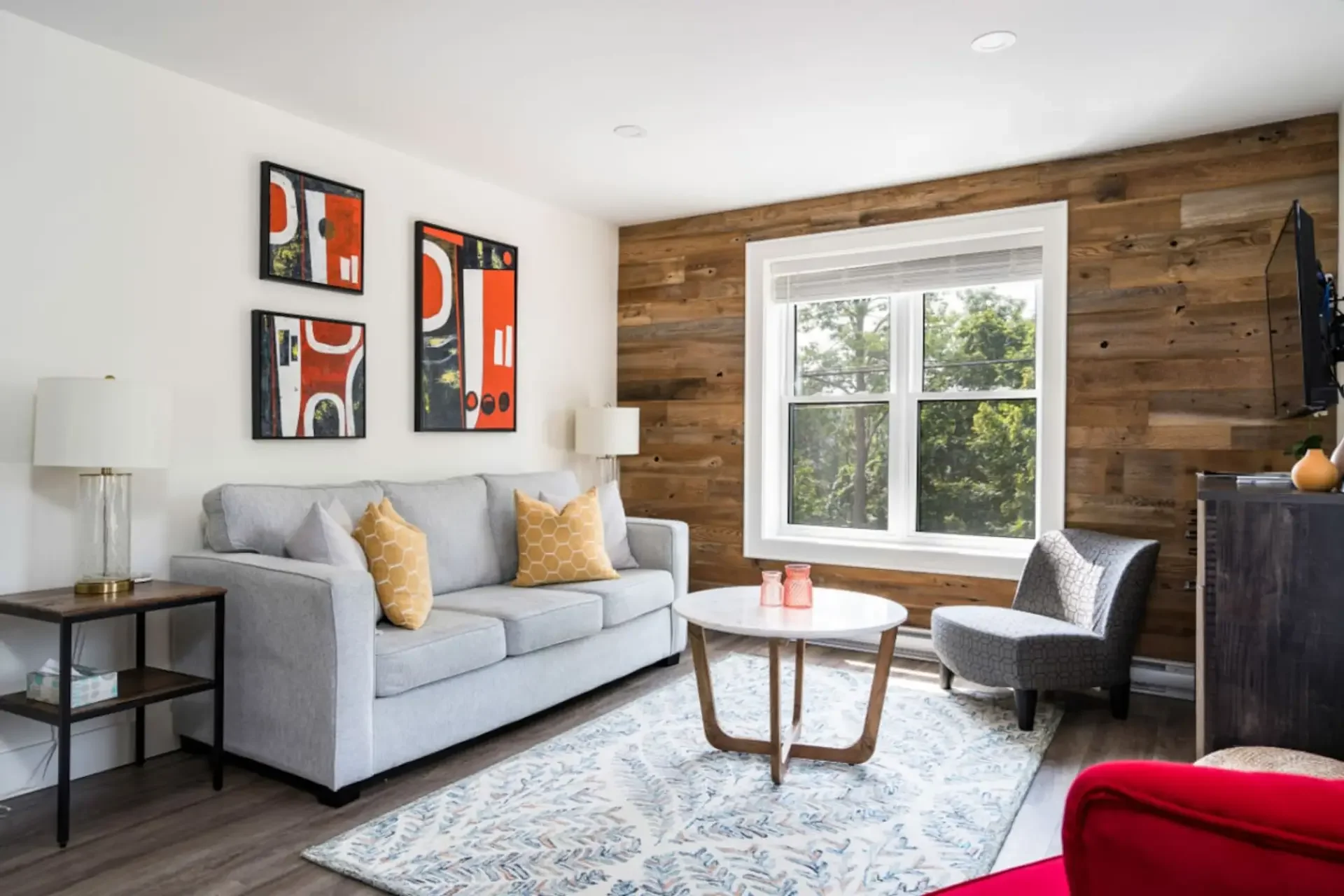 Living room with white walls, wooden accent wall, large window, light gray sofa with yellow patterned pillows, black side table with white lamp, white round coffee table with pink candles, patterned armchair, area rug, abstract art prints on wall, TV on wall, potted plant on cabinet.