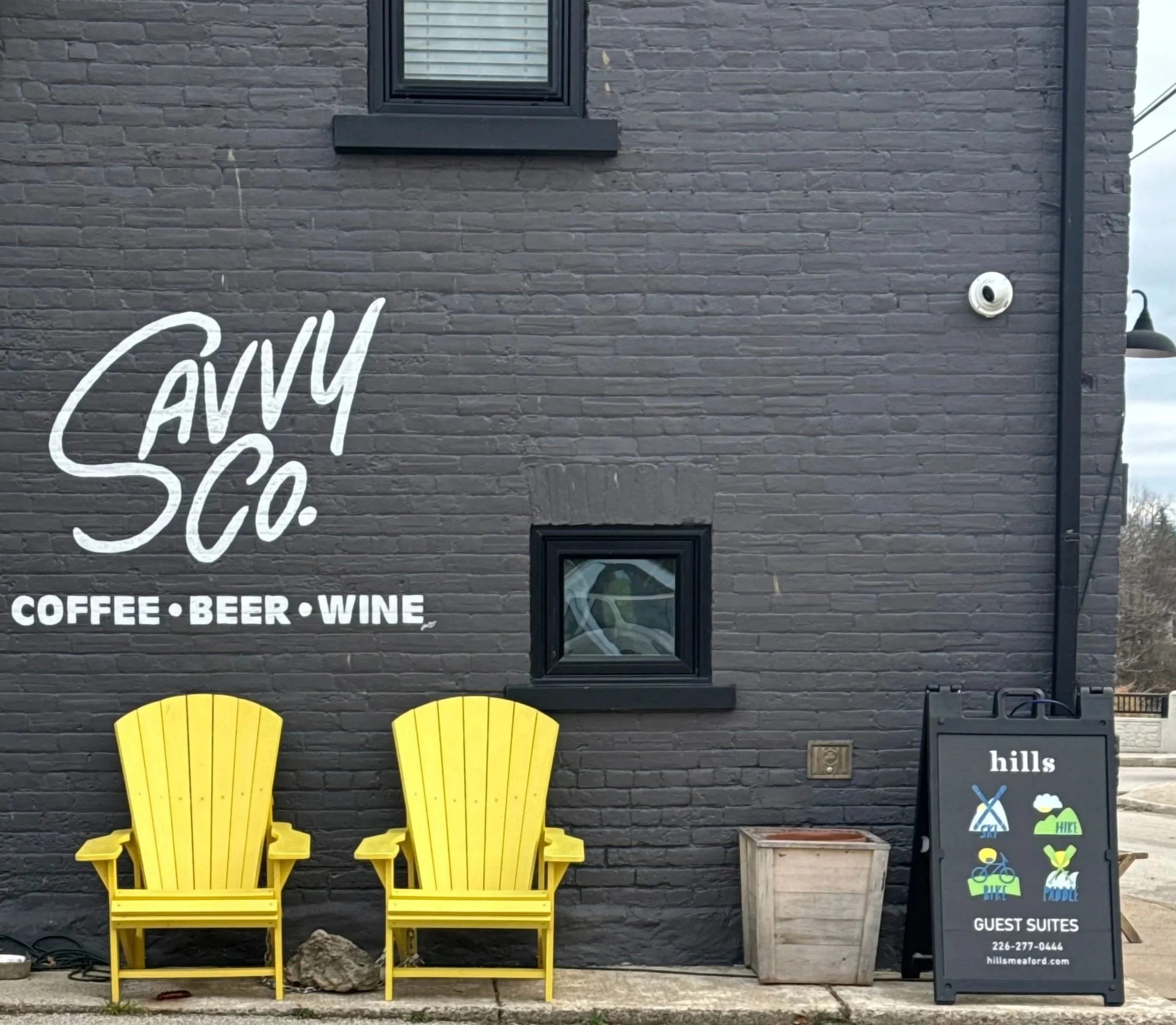 Two bright yellow Adirondack chairs side by side in front of a black brick wall. To the right, a black sign with business information and various symbols for services offered. A small potted plant on the ground next to the sign.