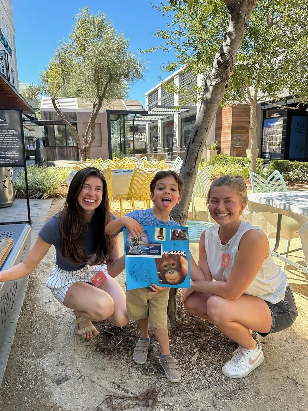 Three people crouching by a tree outside, holding a blue poster with animal images, in a lively outdoor shopping area with yellow and white chairs and greenery.