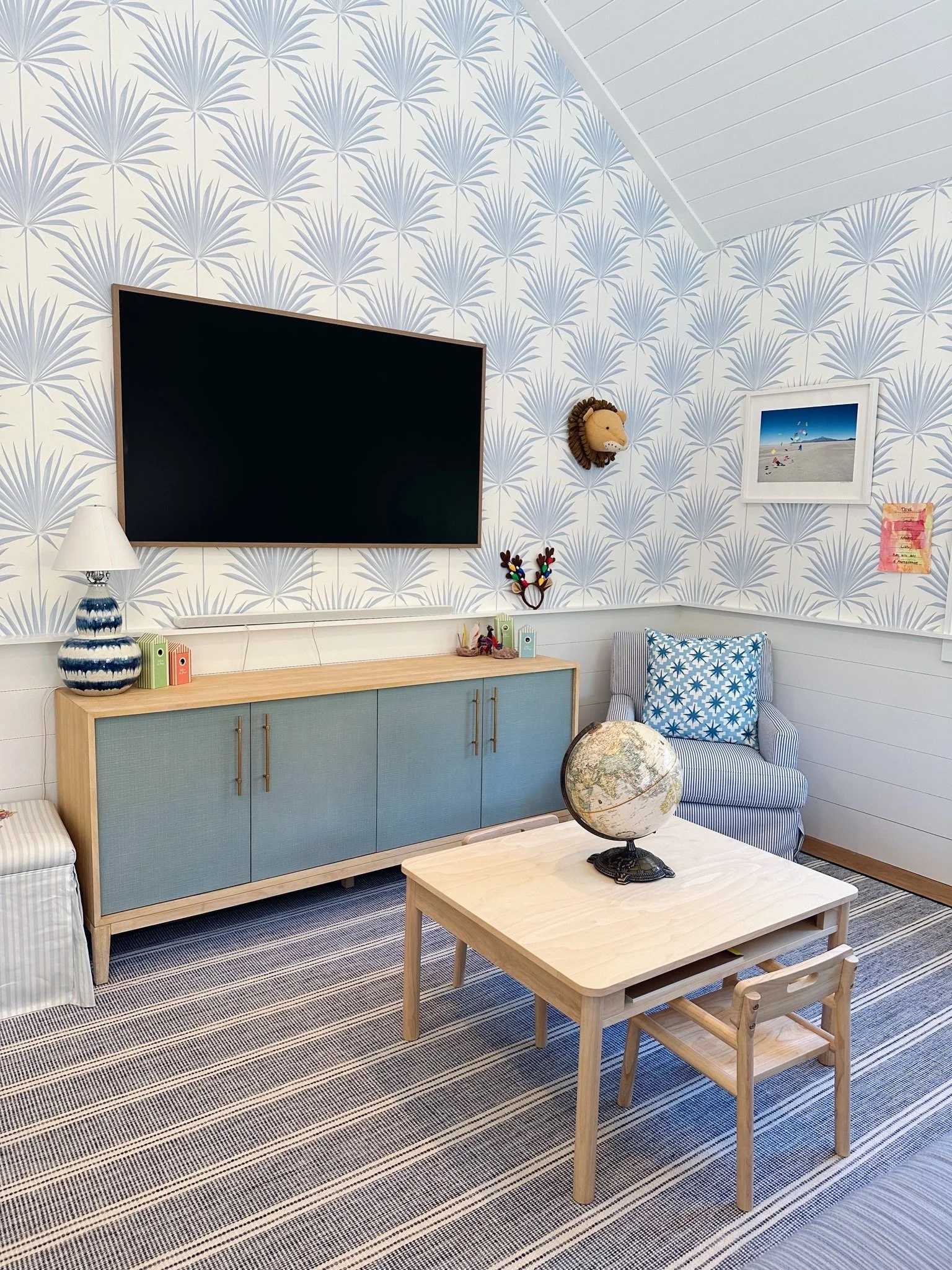 A living room with palm leaf patterned wallpaper, a large flat-screen TV on the wall, a wooden sideboard with blue cabinet doors, a white lamp with a striped blue base, a wall-mounted lion head decor, a framed beach photograph, a colorful reindeer wall decoration, a blue and white patterned pillow on an armchair, a small wooden table with a globe, and a striped blue and white rug.