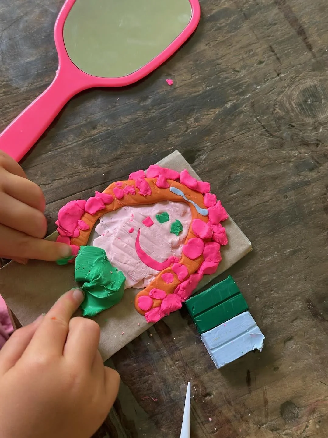 Child making a colorful unicorn craft with playdough, on a wooden table with a pink mirror nearby.