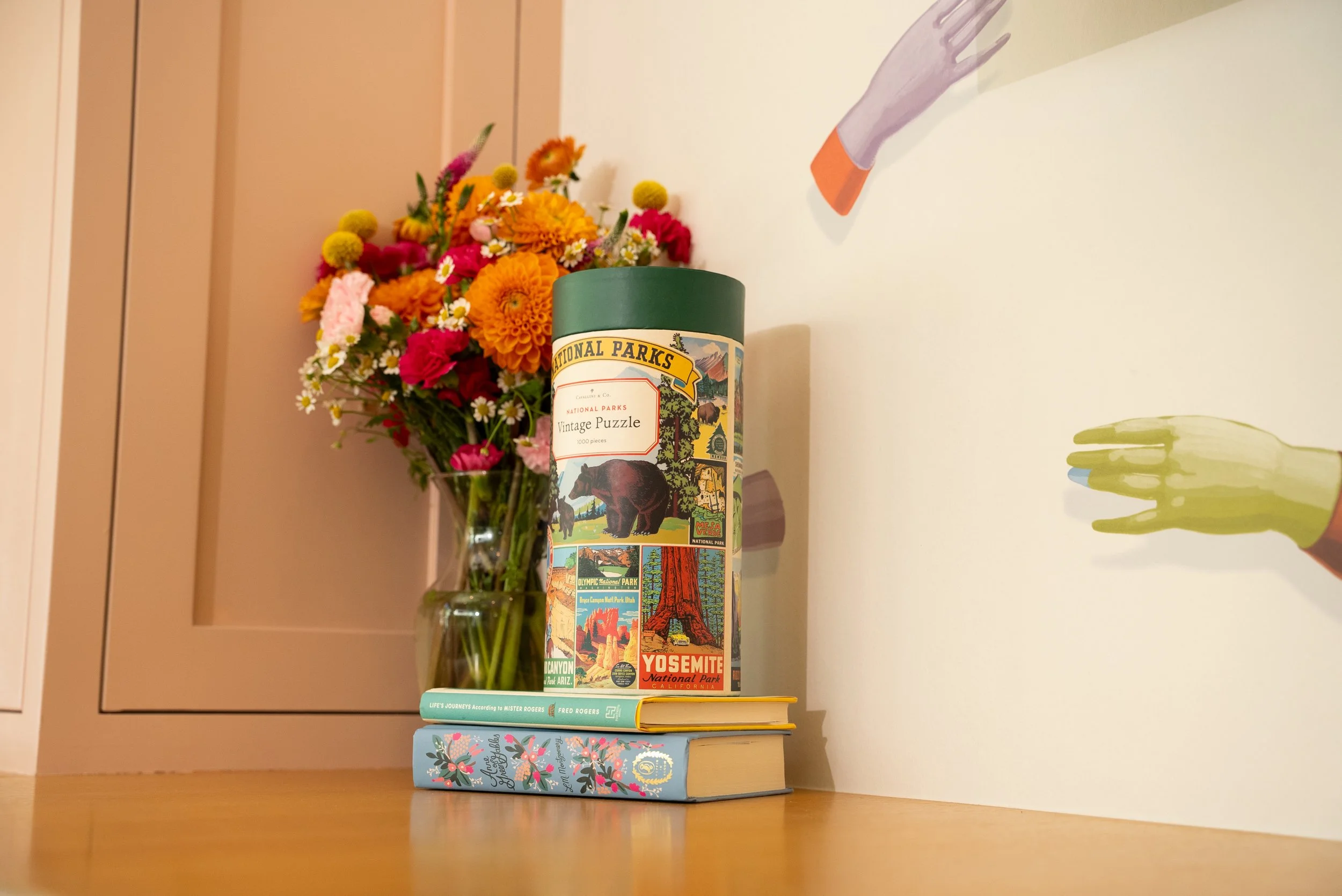 A small glass vase filled with colorful flowers next to a cylindrical puzzle box with vintage park illustrations, placed on two stacked books on a wooden surface against a wall with partial artwork.