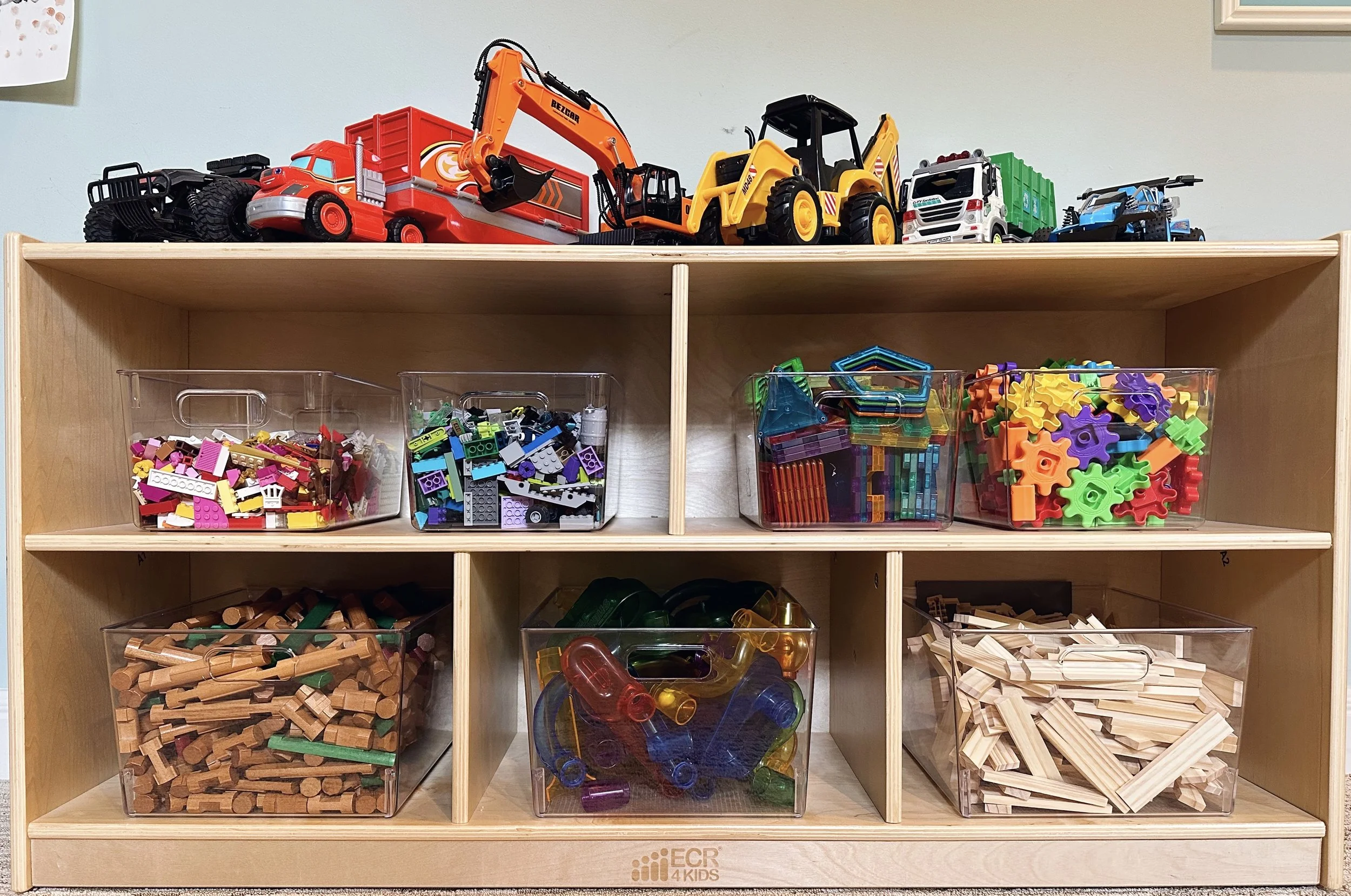 Top shelf with toy construction vehicles including a red fire truck, orange excavator, yellow bulldozer, white dump truck, and blue motorcycle; middle and bottom shelves with clear plastic bins containing assorted building blocks, gears, interlocking pieces, and wooden construction pieces.