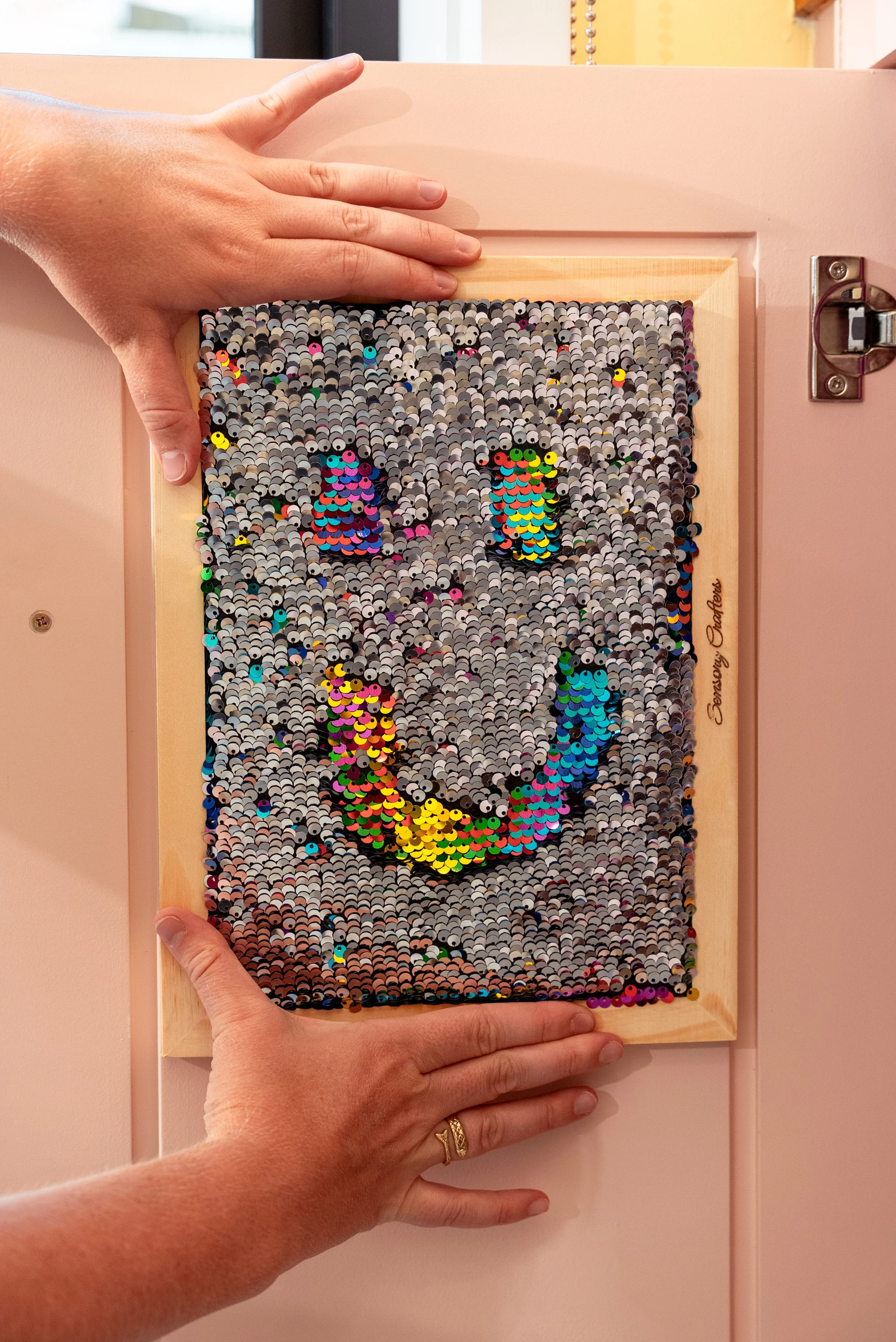 Sequin mosaic of a smiling face with colorful details, mounted on a pink wall with hands holding the frame.