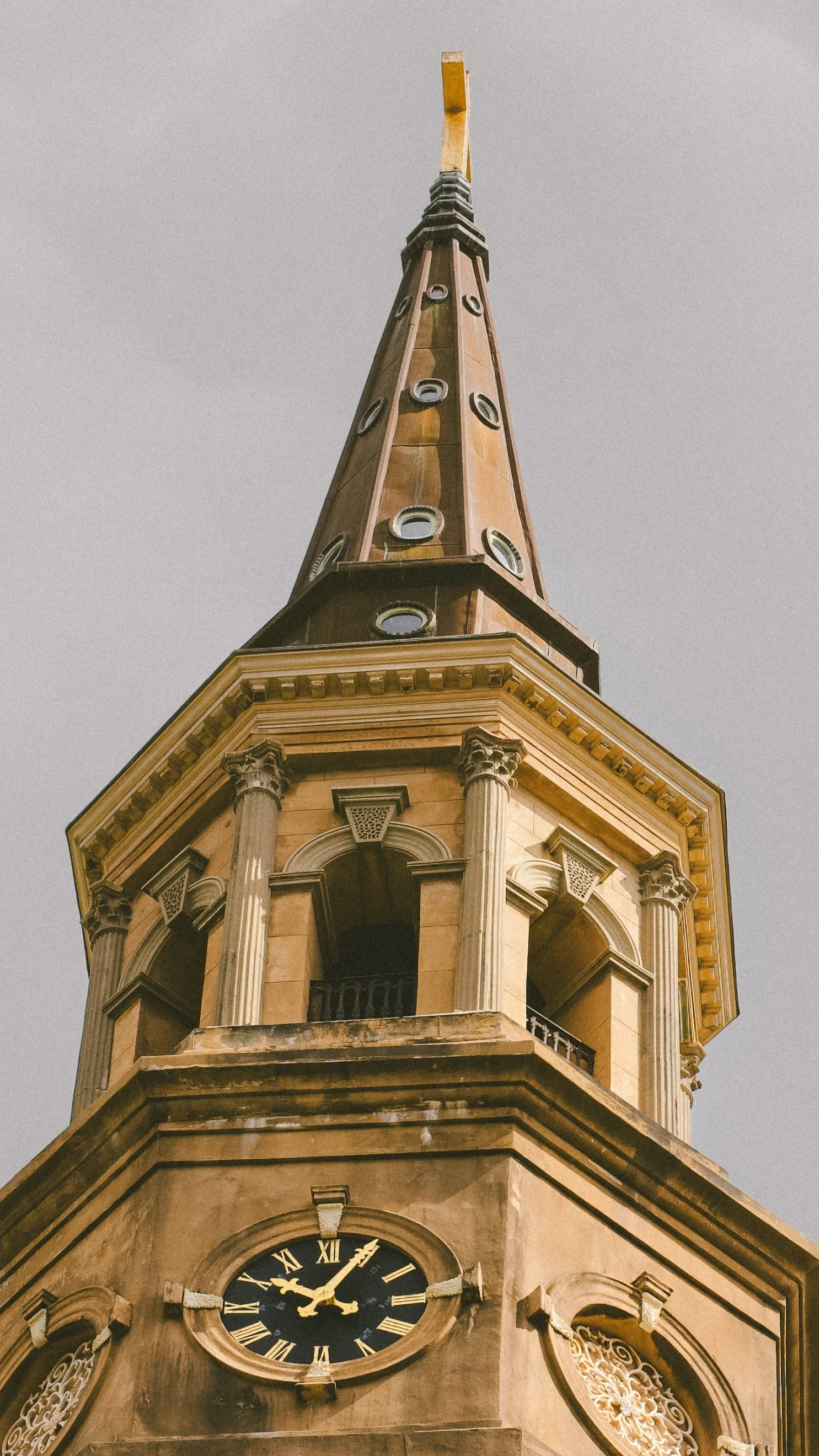 Close-up view of a historic church steeple with a clock, featuring classical architectural elements including columns, arched windows, and decorative details, against a cloudy sky.
