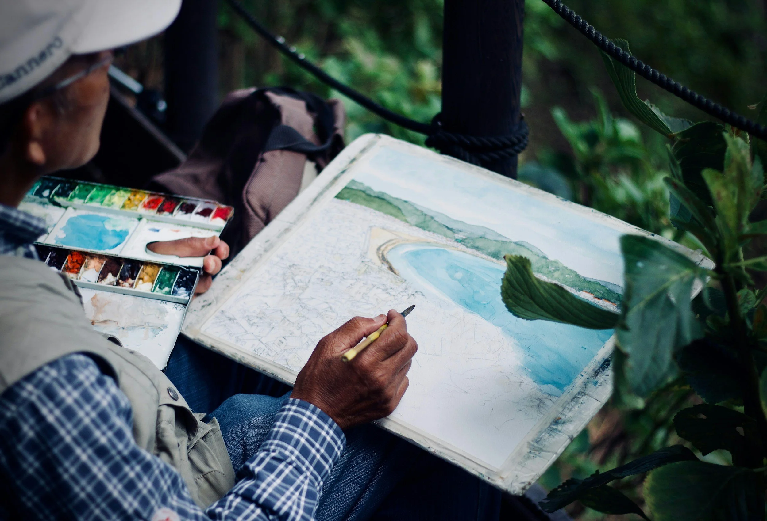 An artist painting a landscape scene with watercolors on a palette, depicting a lake, green hills, and a cloudy sky, sitting outdoors among greenery.