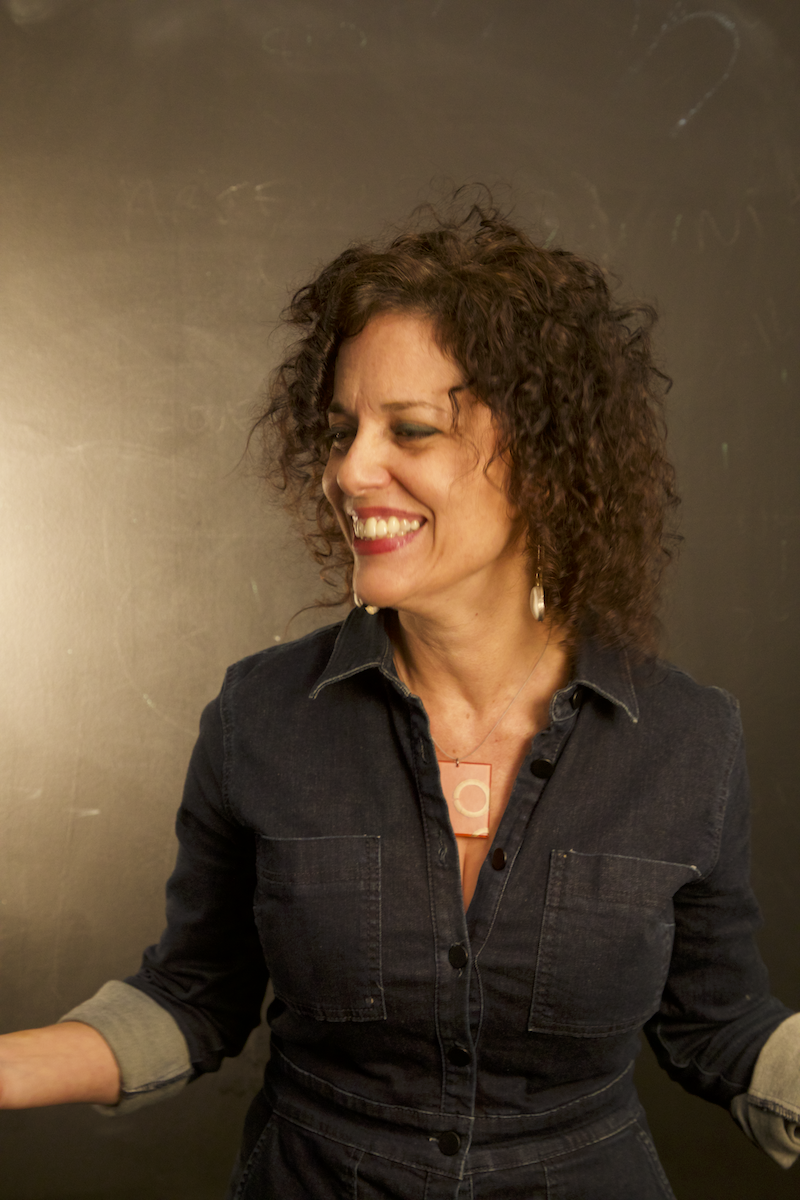 A woman with curly brown hair wearing a black denim shirt laughing with her head tilted to the side against a dark background.