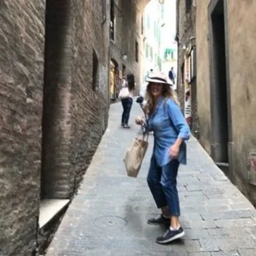 Woman in a narrow stone alley wearing a hat and denim outfit, holding a bag, with another person further up the alley.