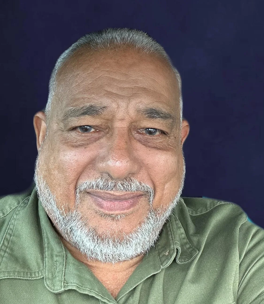 Primer plano de un hombre mayor sonriente con pelo gris, barba y ojos azules, vestido con una camisa verde sobre un fondo oscuro.