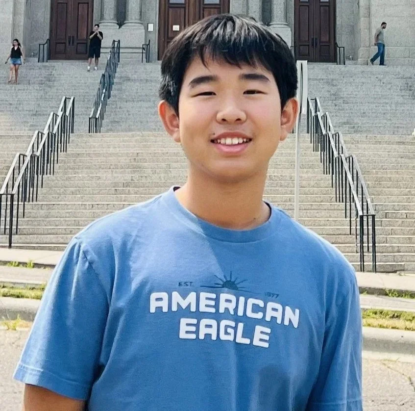 Close-up of a young boy with short black hair, smiling slightly, wearing a black collared shirt with red trim.