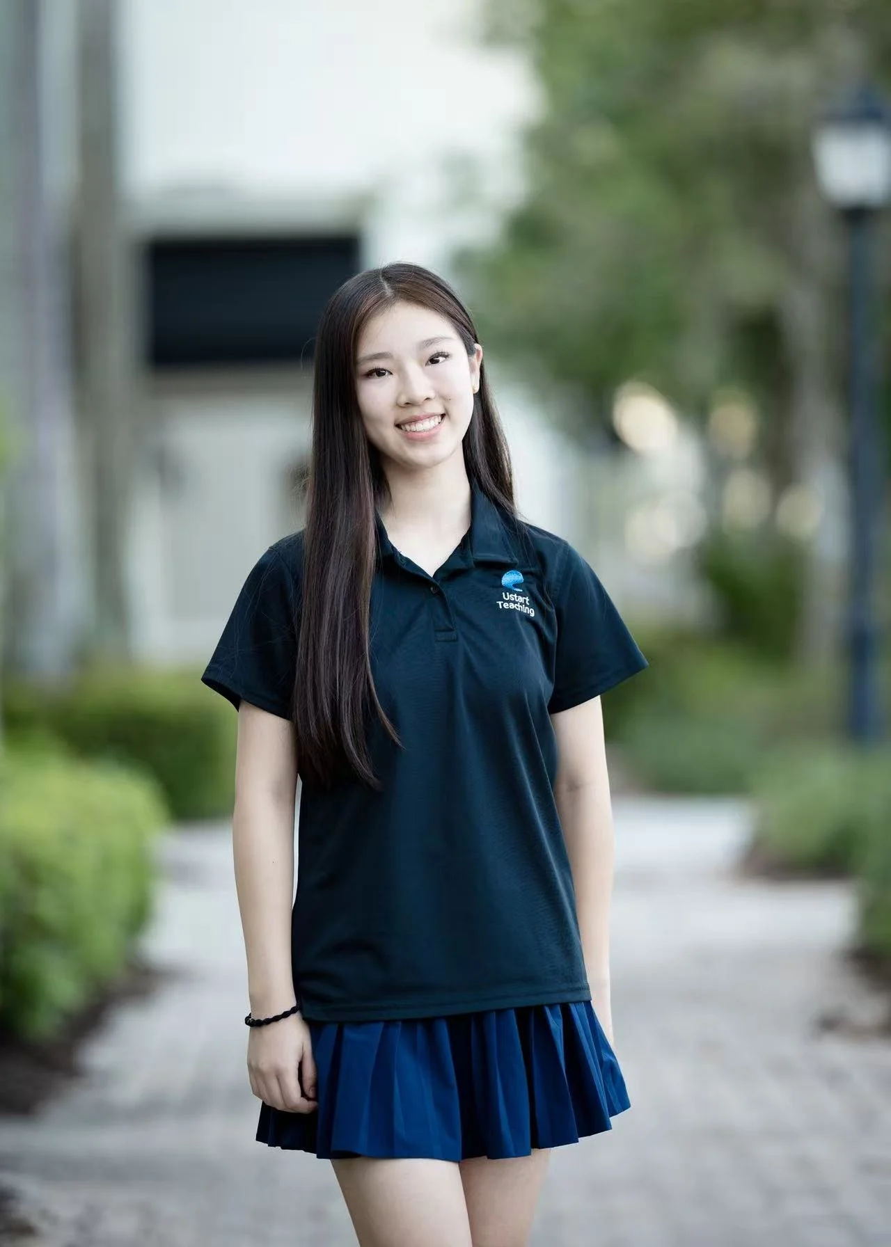A young woman with shoulder-length brown hair and wearing a white shirt, standing outdoors with green foliage in the background.