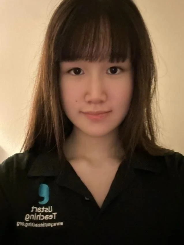 Young man standing outdoors in front of pink flowers and a brick wall, wearing a black polo shirt with a logo on it.