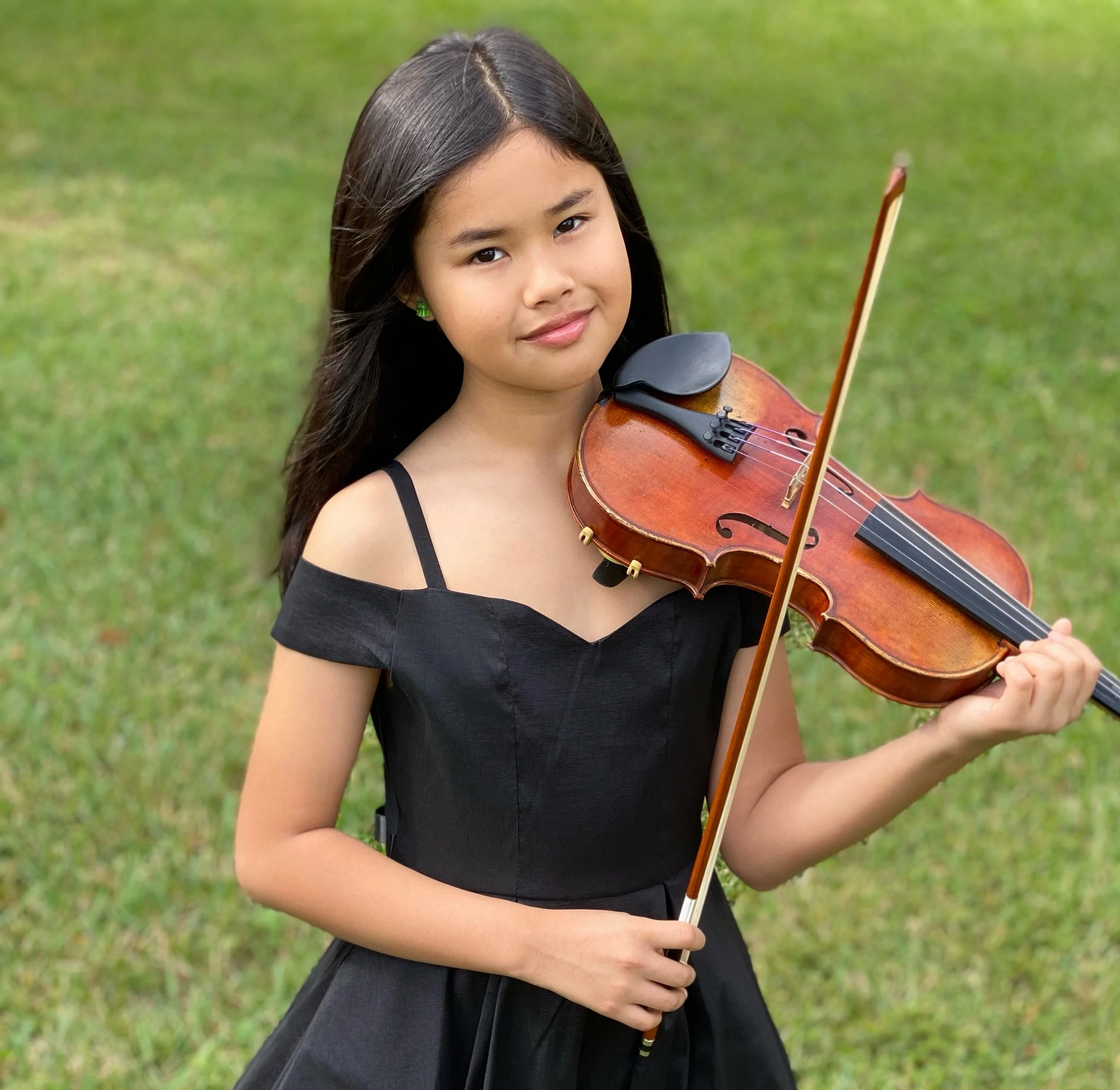 A young girl in a black dress holding a violin and bow outdoors on green grass.