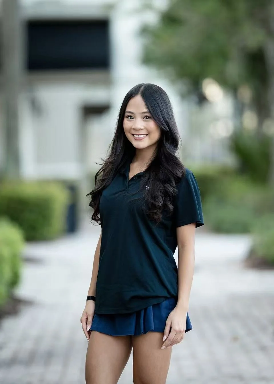 Portrait of a young woman with long dark hair wearing a green shirt standing against a white textured wall.