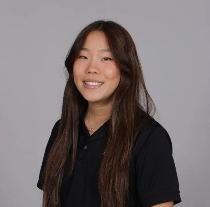 Portrait of a young woman with long dark hair wearing a green shirt standing against a white textured wall.