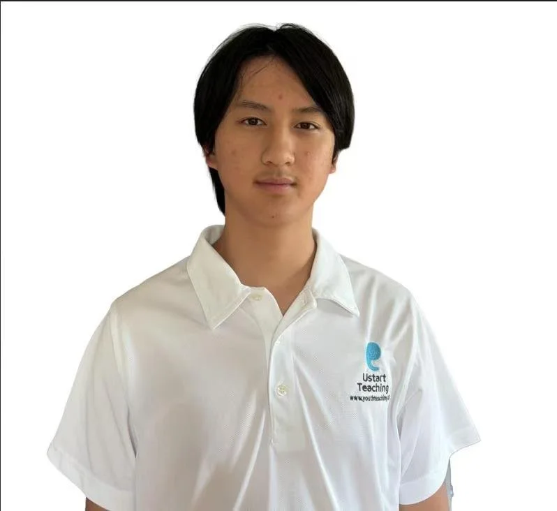 A young woman with long black hair wearing a white collared shirt standing against a plain light-colored wall.