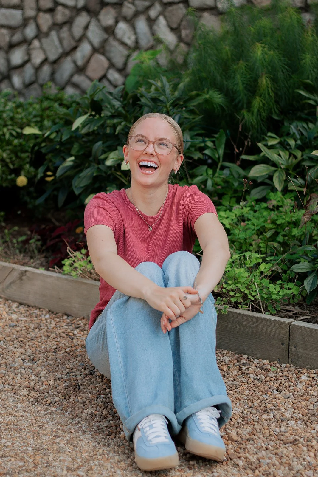 A woman sitting on the ground, laughing, in a garden with green bushes and a stone wall in the background.