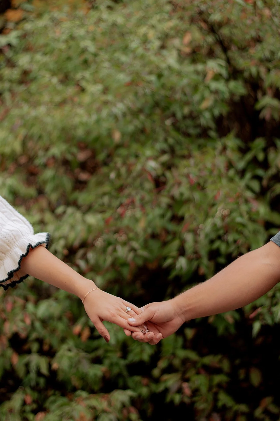 Two people, one wearing a white sleeve and the other in a darker sleeve, hold hands against a background of green bushes.