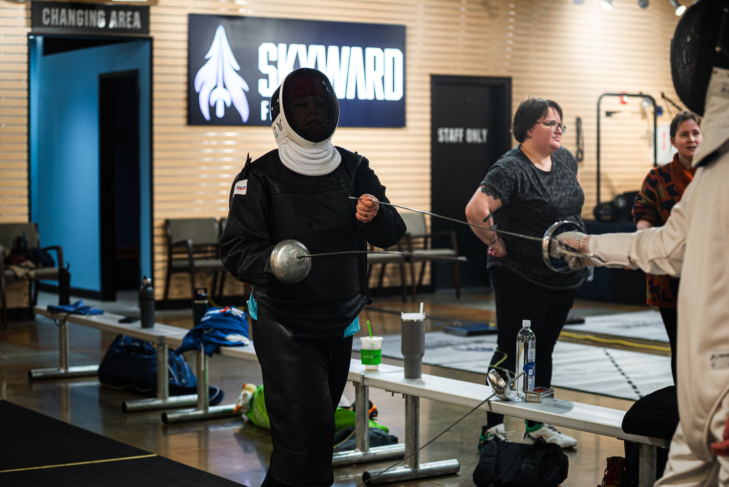 A person in fencing gear standing in an indoor fencing facility, holding a fencing epee, with others and various fencing equipment in the background.