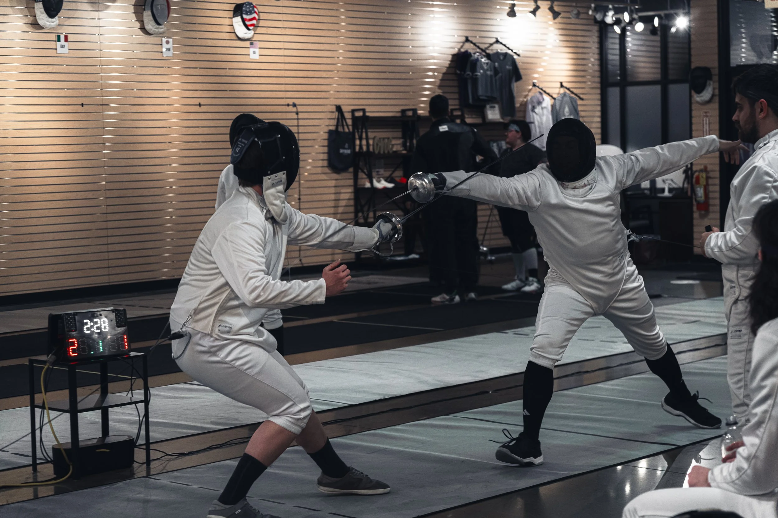 Two fencers competing in a fencing match indoors, wearing protective gear and masks, with a scoreboard nearby, and other people in the background.