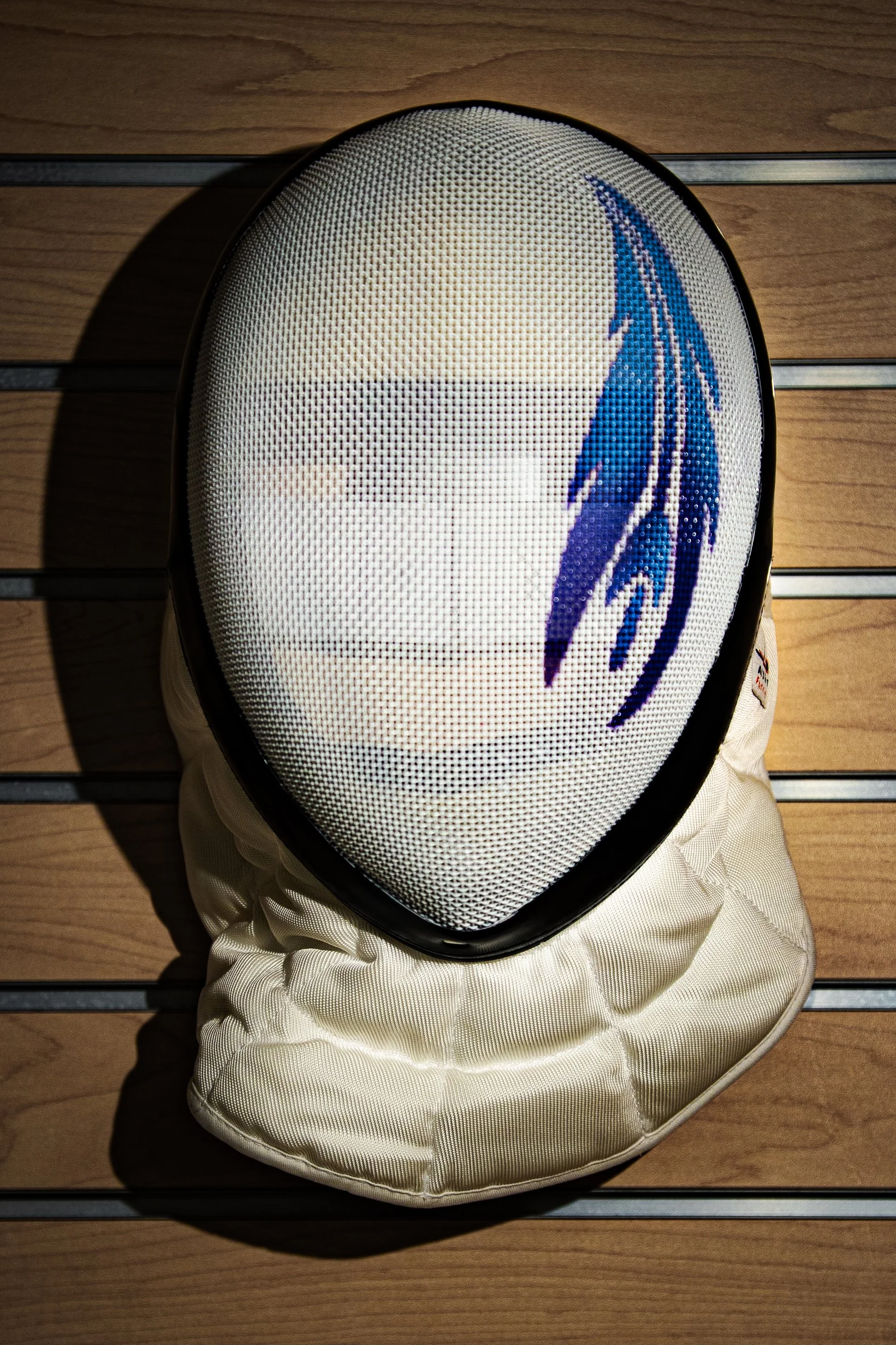 A white fencing mask with a custom skyward fencing logo design on the front, resting on a wooden surface.