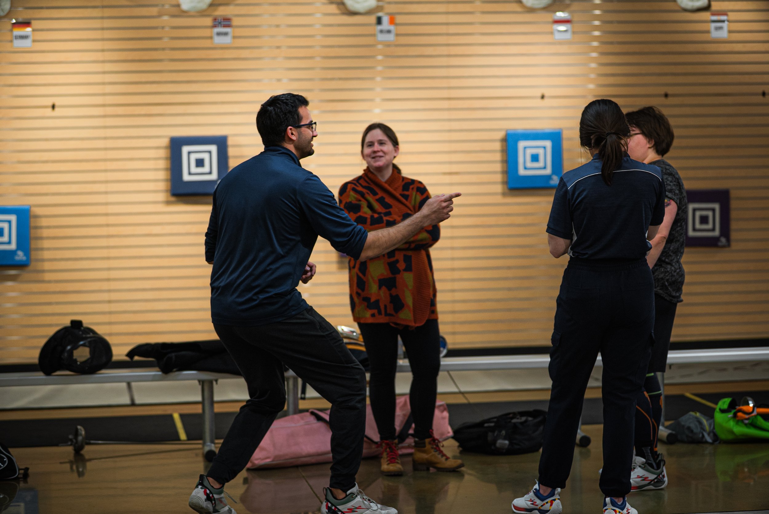 Four people in a gym or sports facility engaging in conversation. One person is dancing, and others are standing nearby with sports gear around them.